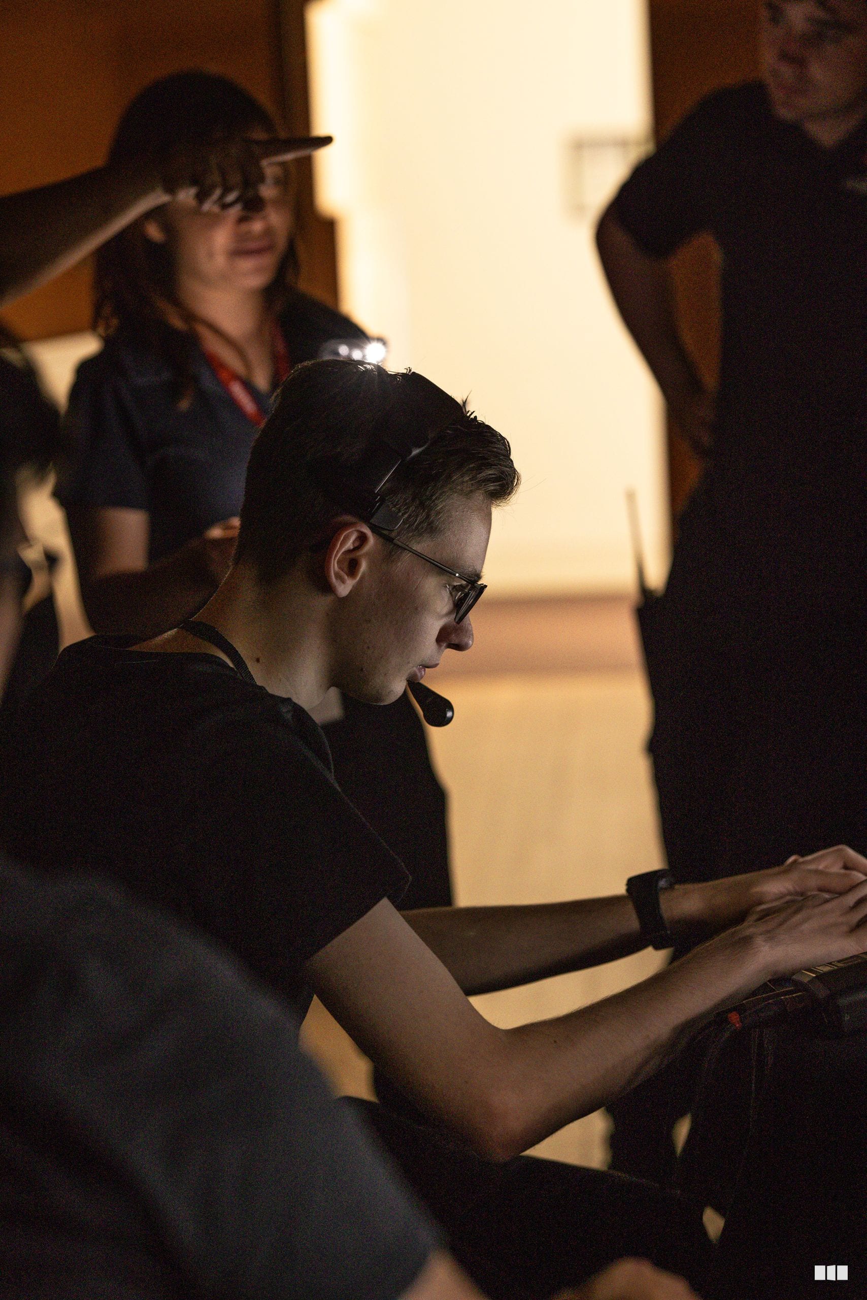 A musician wearing glasses focuses intently while playing a keyboard instrument during a rehearsal or performance with ensemble members visible in the background.