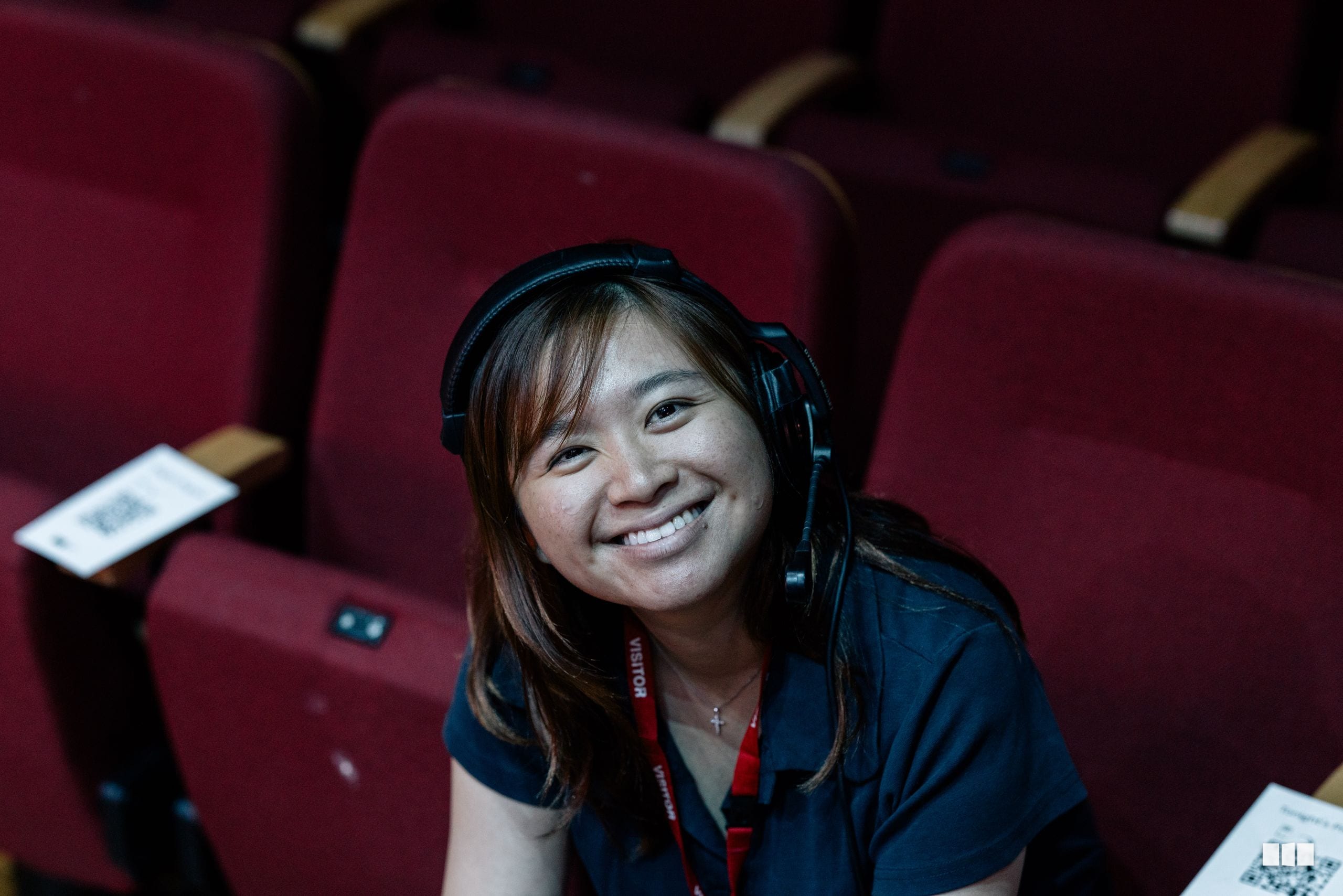 A young woman wearing headphones and a credential lanyard smiles while seated in a theater with burgundy seats.