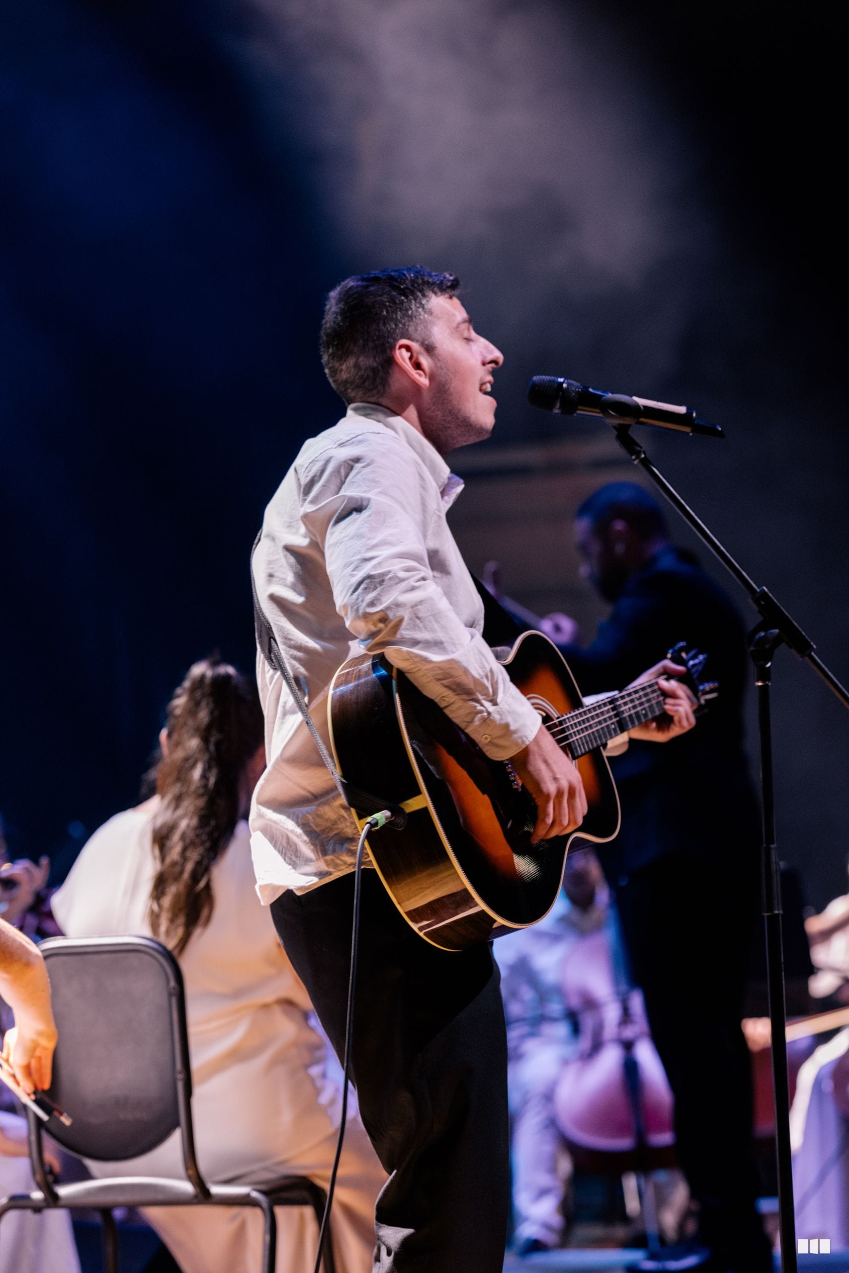 A male soloist performs with an acoustic guitar at the microphone during a concert performance.