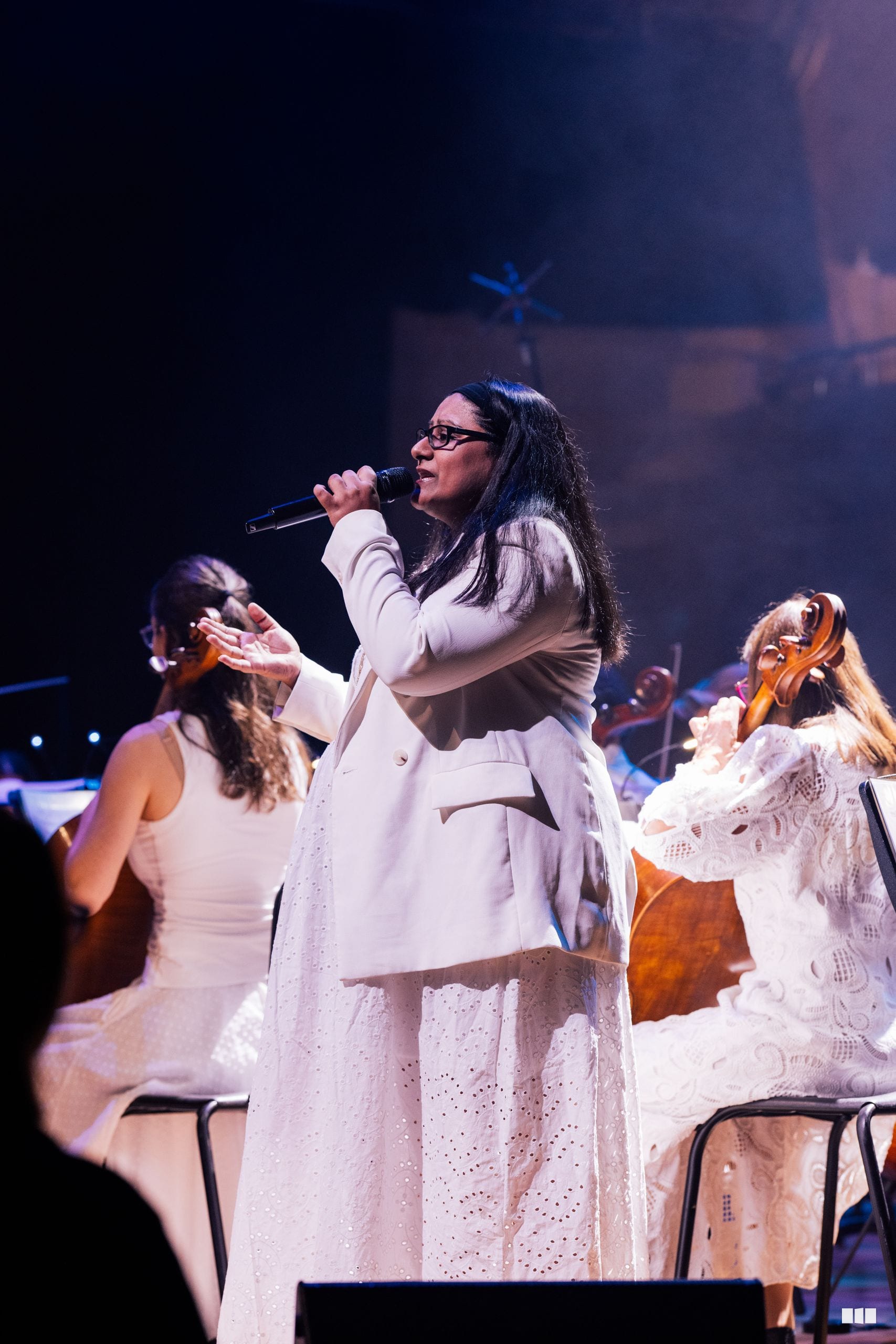 A soloist in white attire performs with a microphone while accompanied by string musicians in an orchestral setting.