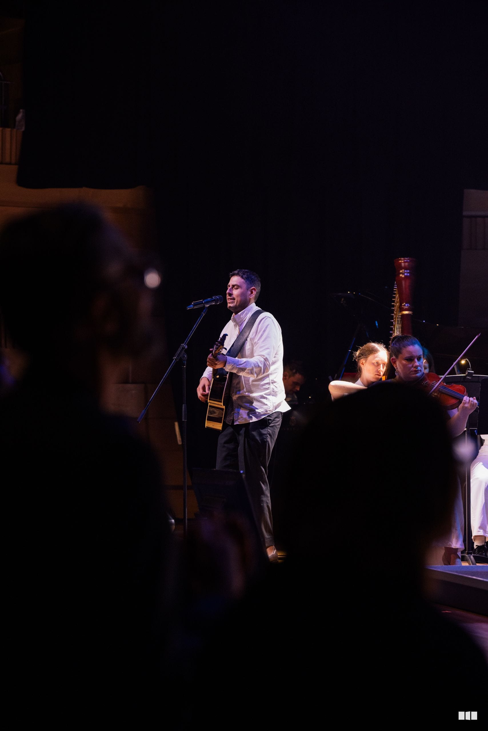 A male soloist performs with an acoustic guitar at the microphone while string musicians accompany him on stage.