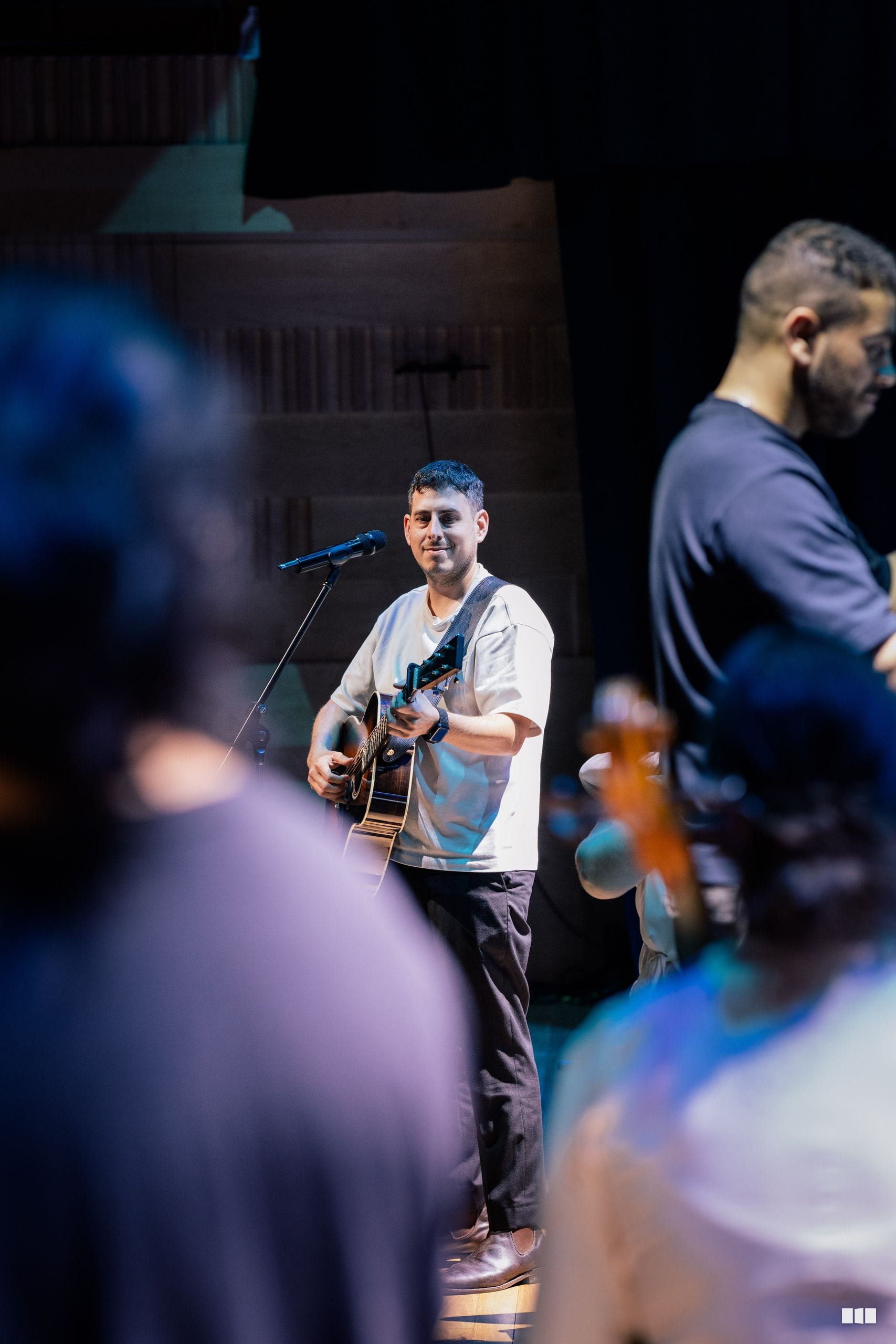 A guitarist performing solo on stage with a microphone while audience members watch from the foreground.