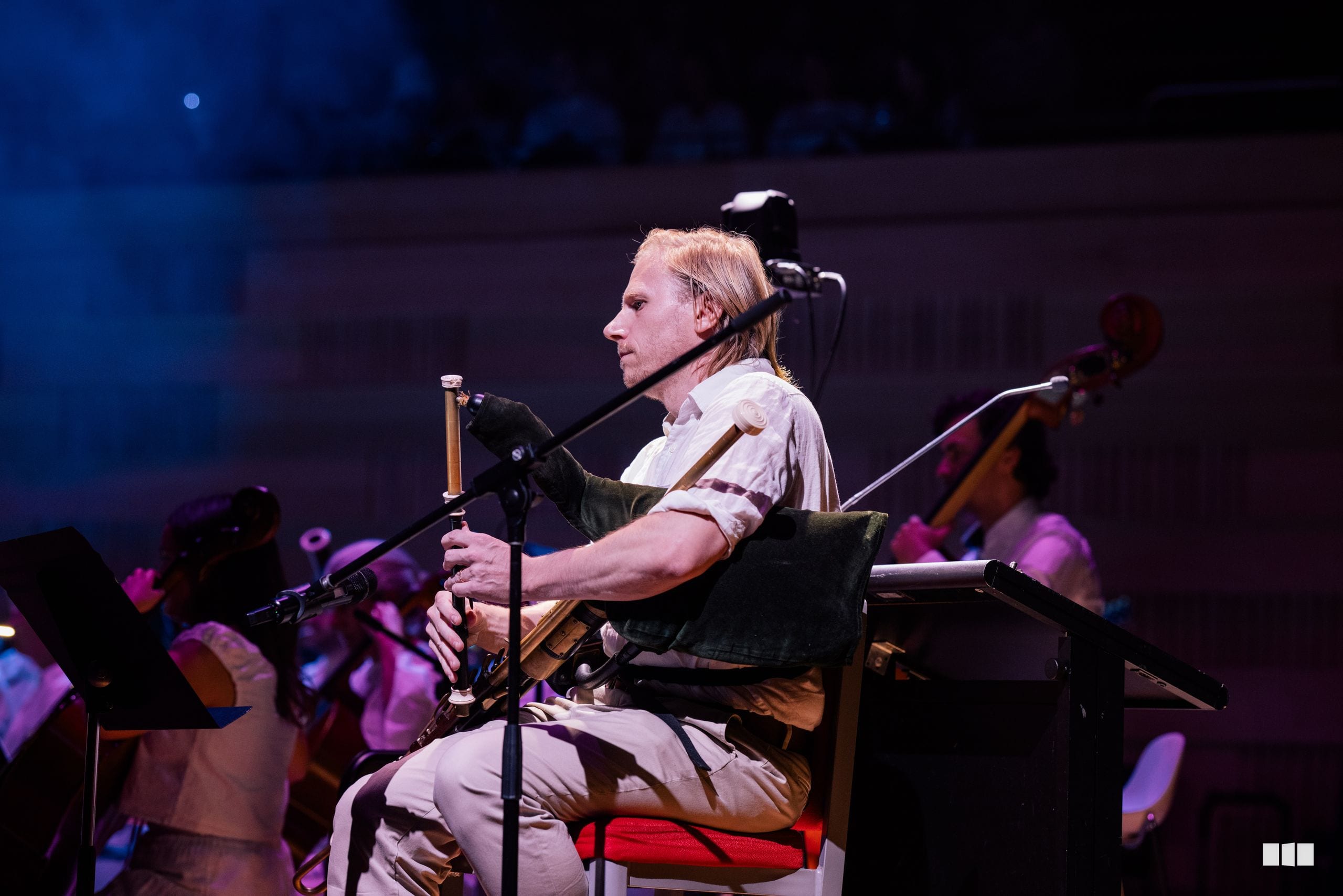 A musician plays a stringed instrument while seated on stage, accompanied by an orchestra in warm stage lighting.