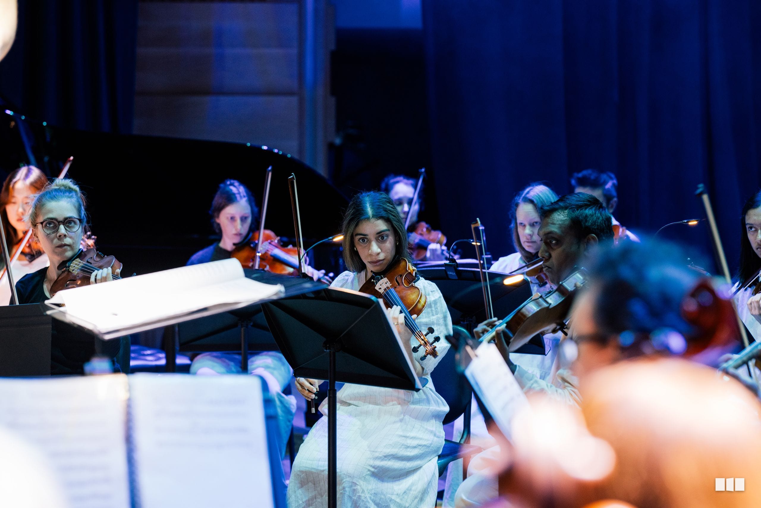 Young violinist in white shirt performs with orchestra members in blue stage lighting during a rehearsal or performance.