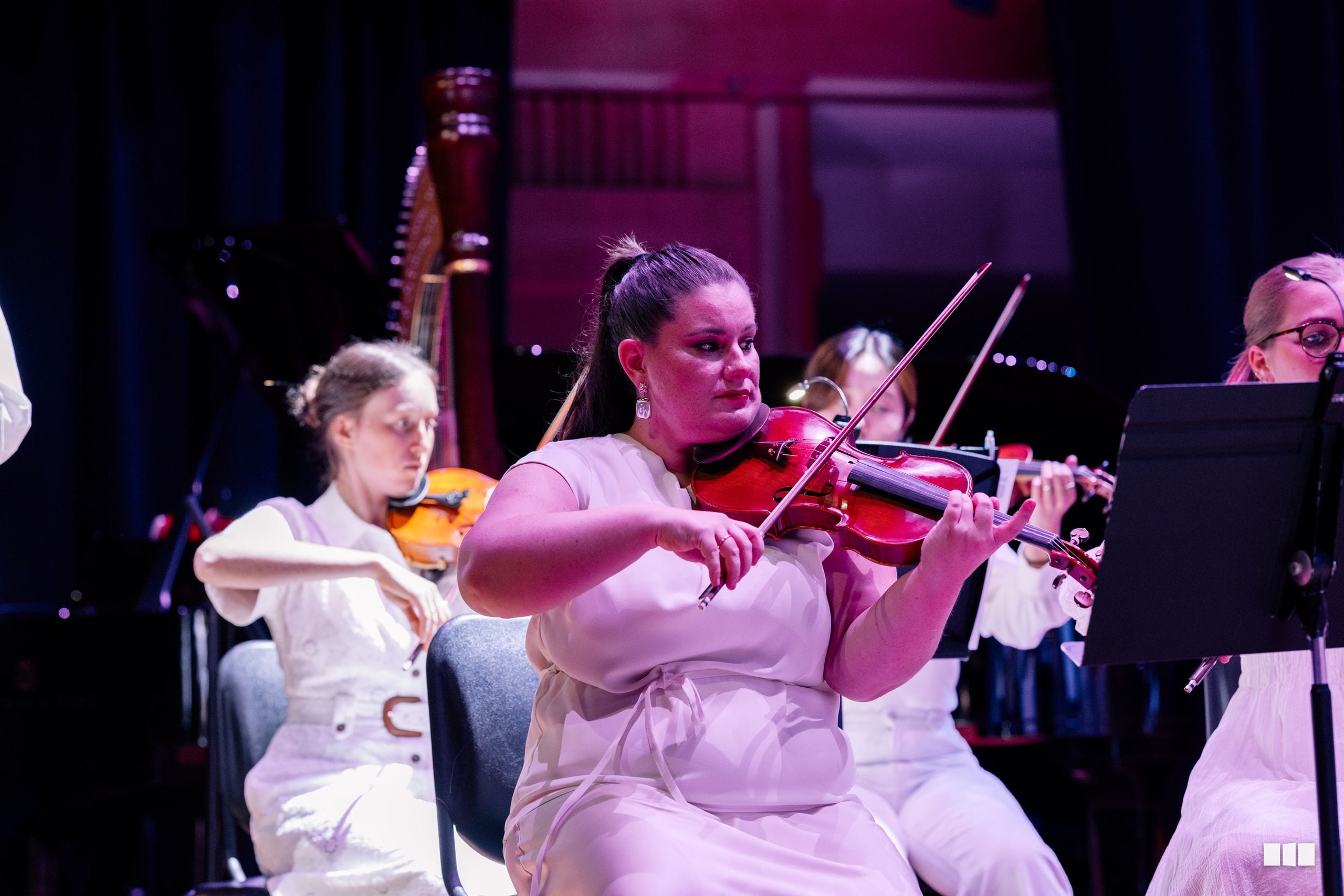 A violinist in a pink dress performs with fellow orchestra members during a concert under warm stage lighting.