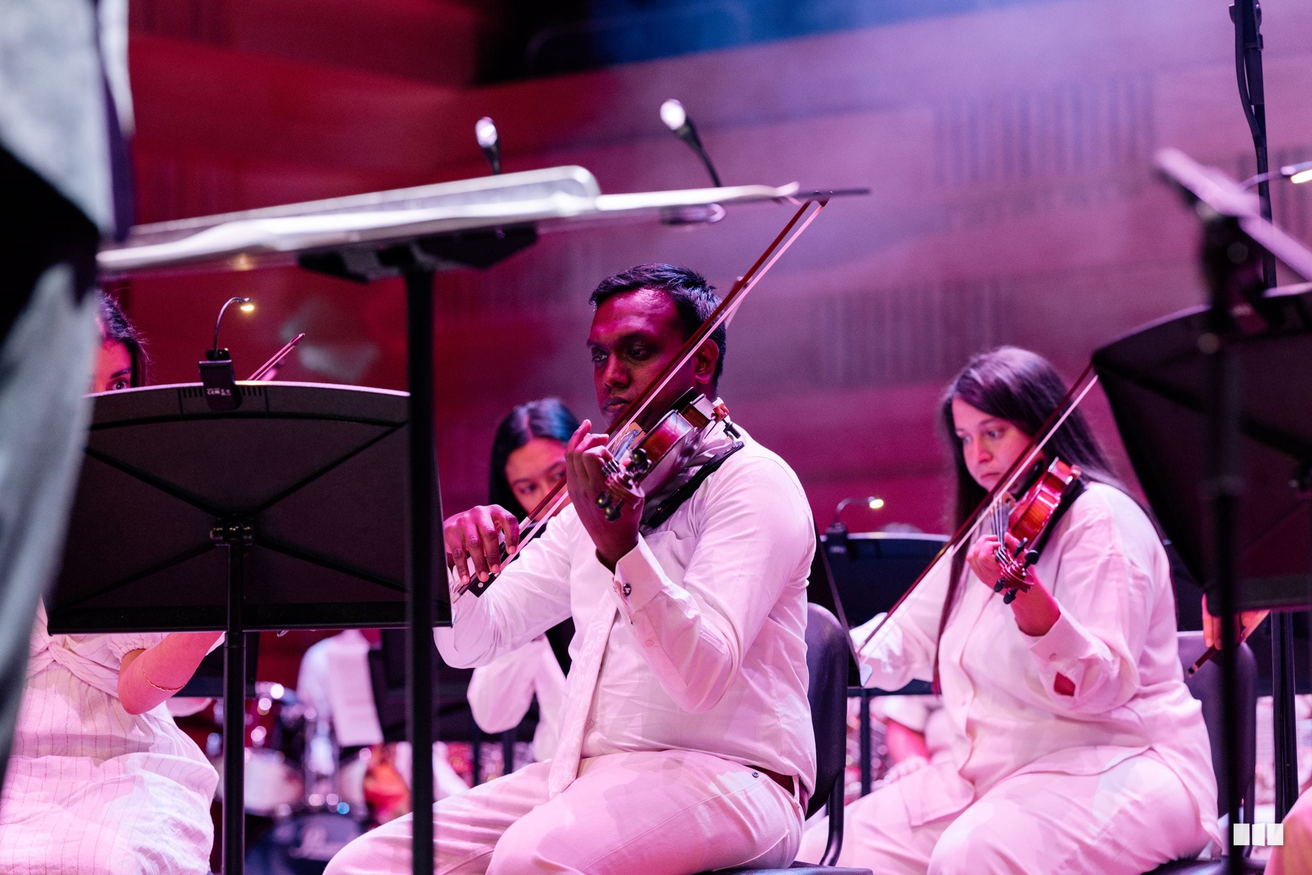Two violinists in white formal attire perform together on stage with pink atmospheric lighting during an orchestral performance.