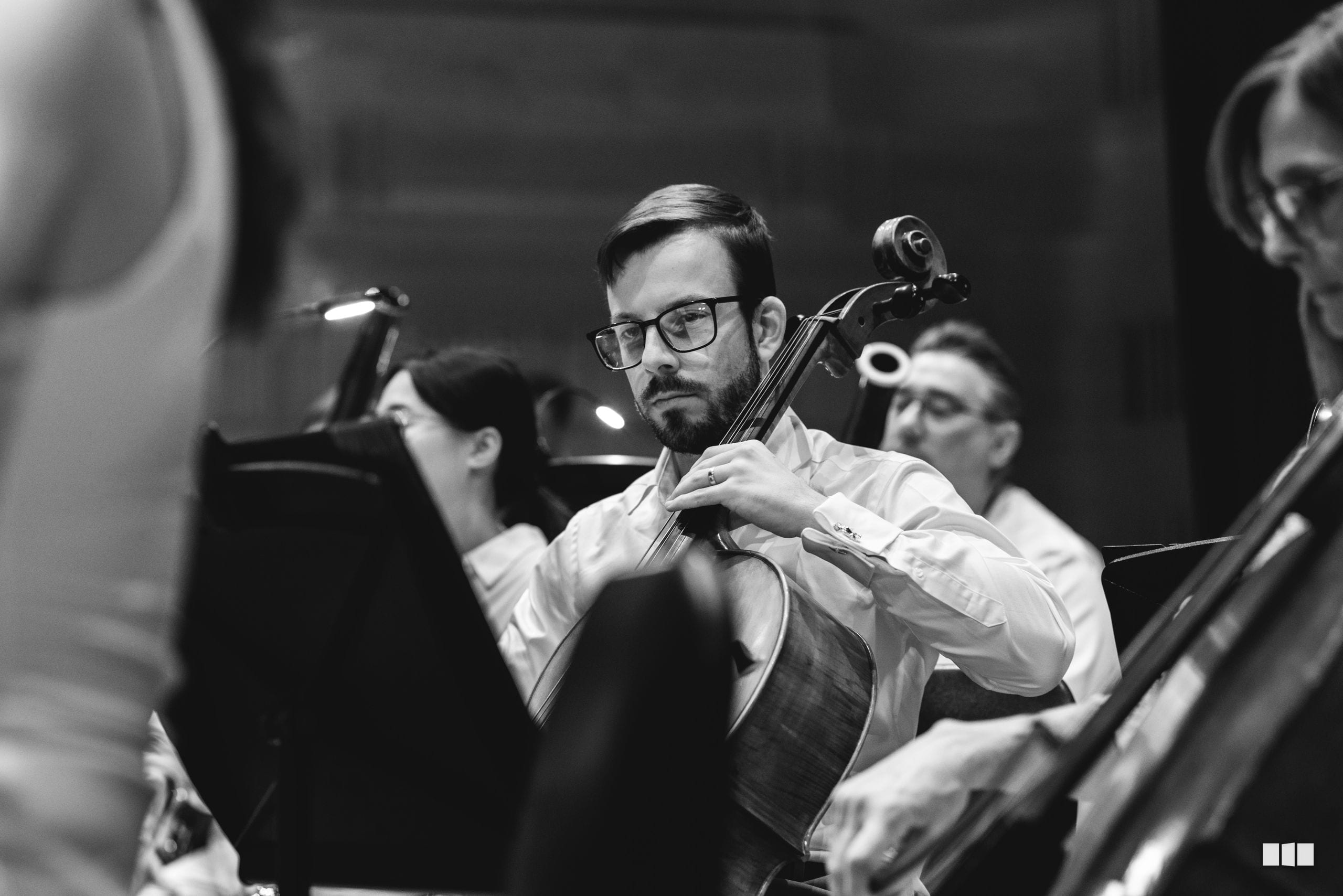 A cellist in formal white shirt performs with concentration during an orchestral rehearsal or recording session.