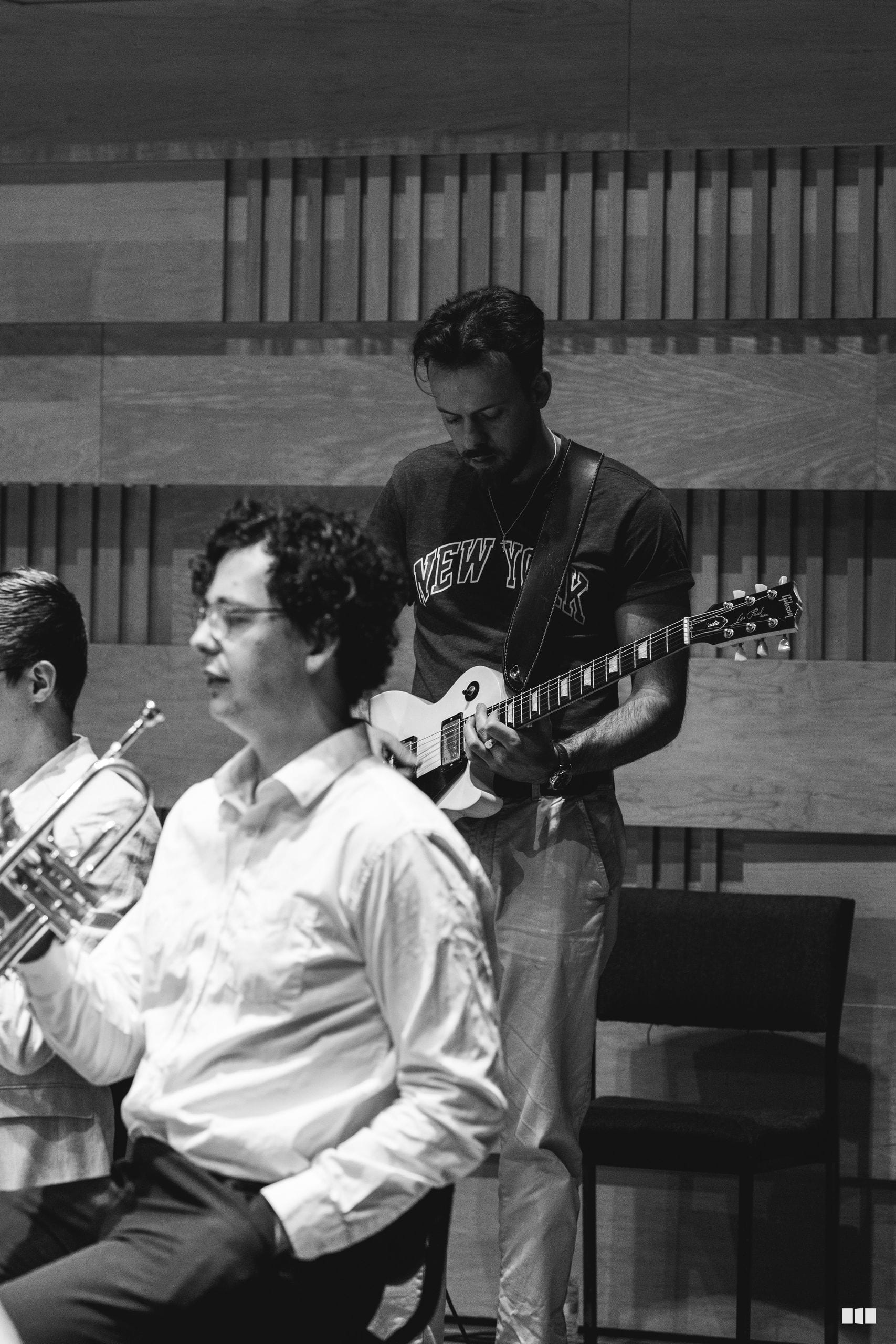 A guitarist in a New York shirt performs alongside brass and wind musicians in a rehearsal or performance setting.
