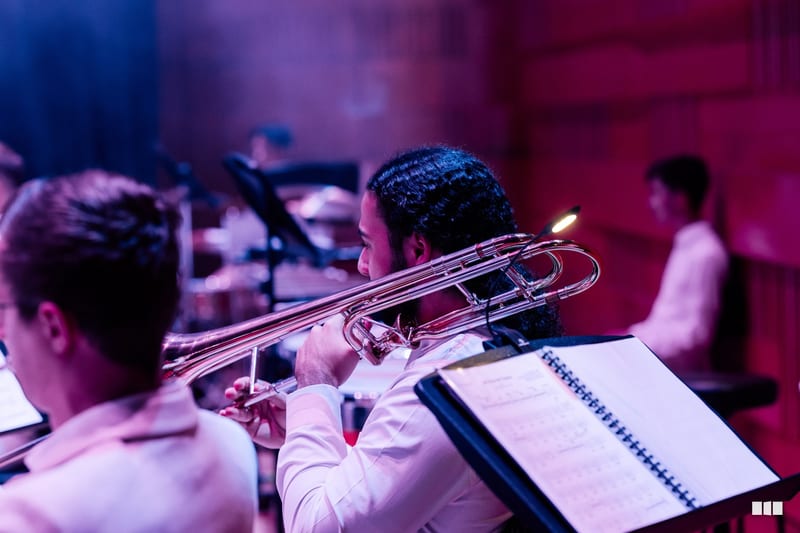 A trombonist in formal attire performs with sheet music during an orchestral rehearsal or concert.