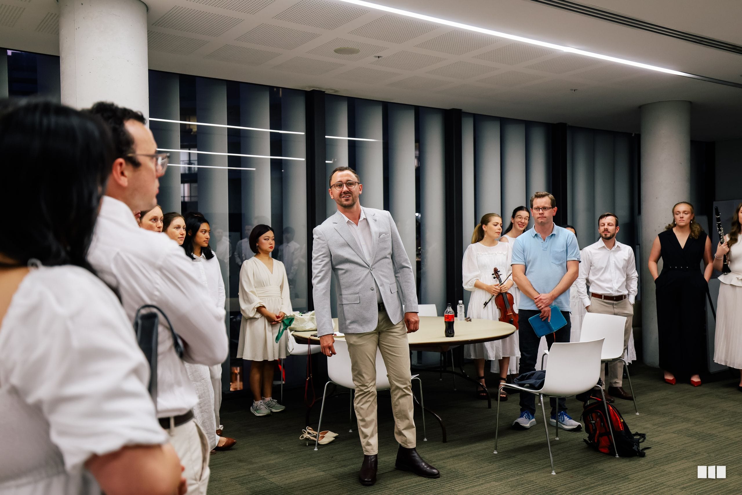 A man in a light gray blazer stands centrally in a modern rehearsal space while musicians and ensemble members gather around him during what appears to be a musical workshop or masterclass.