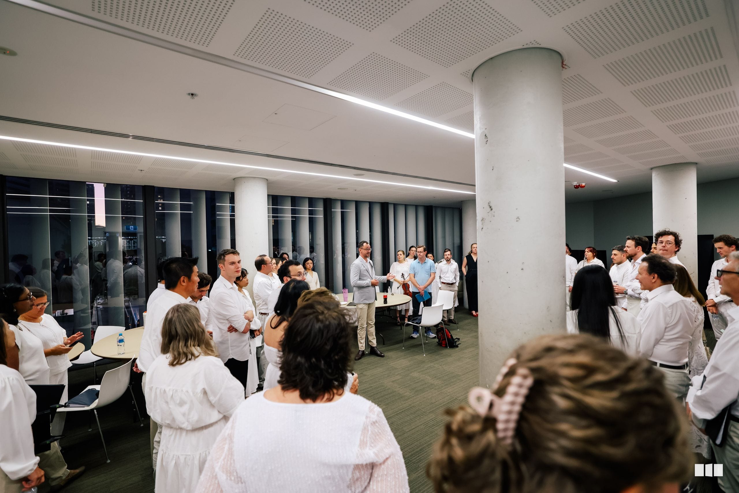 A large group of people wearing white shirts gather in a modern office space with white columns for what appears to be a social or networking event.