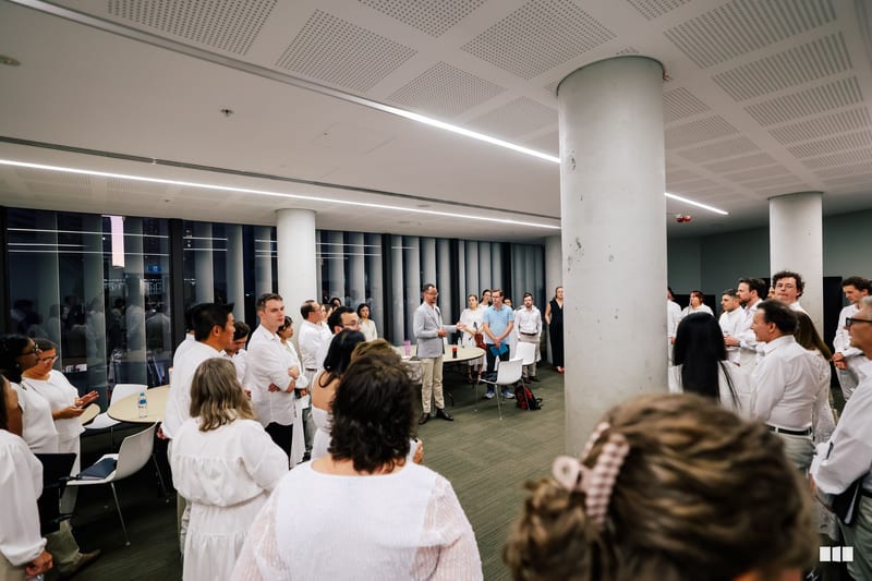 A large group of people wearing white shirts gather in a modern office space with white columns for what appears to be a social or networking event.