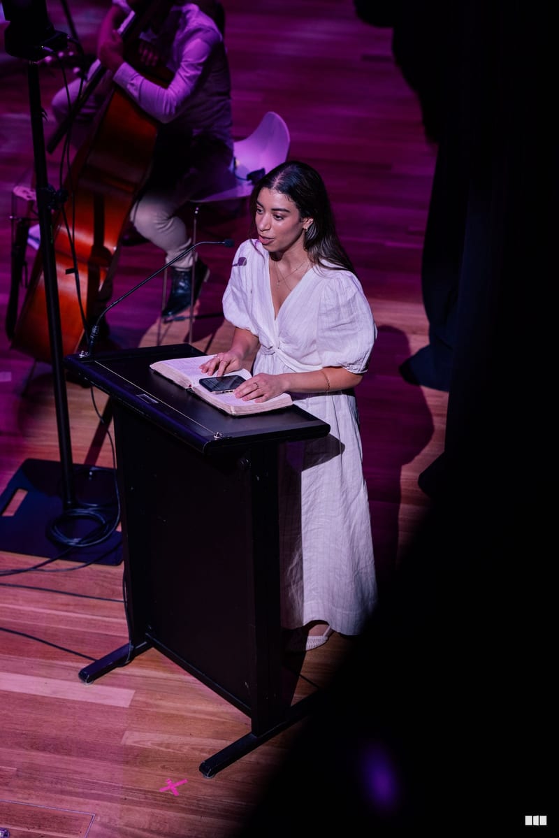 A woman in a white dress reads from a document at a podium during a performance with orchestra musicians and purple stage lighting in the background.