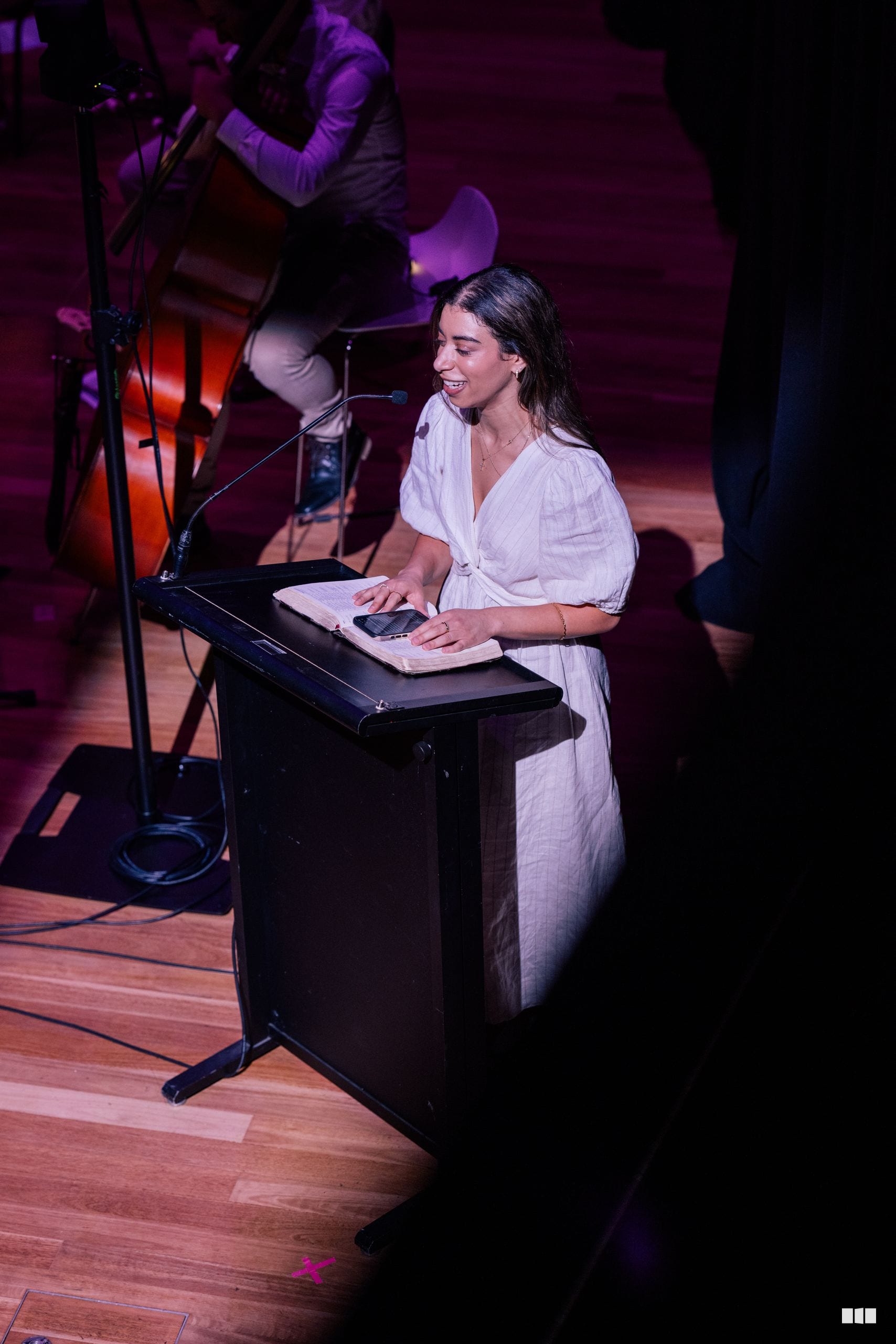 A woman in a white shirt speaks at a podium with a microphone during a performance, with a musician playing cello visible in the background.