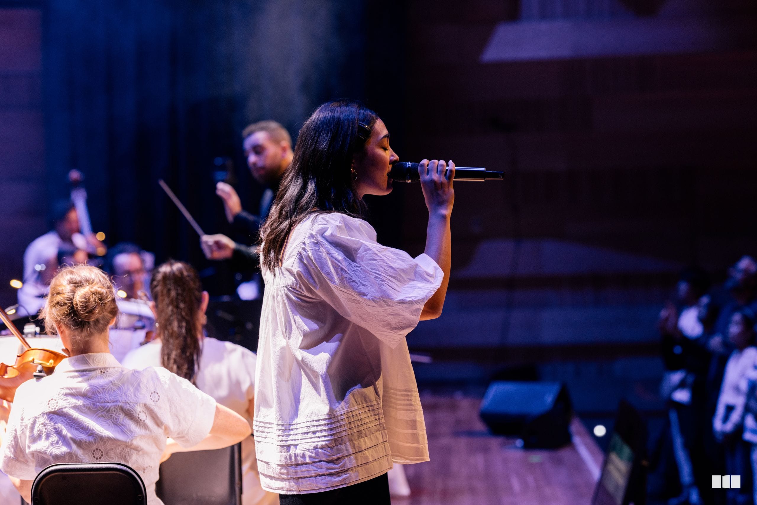 A female soloist performs with microphone while accompanied by orchestra musicians in the background during a concert performance.