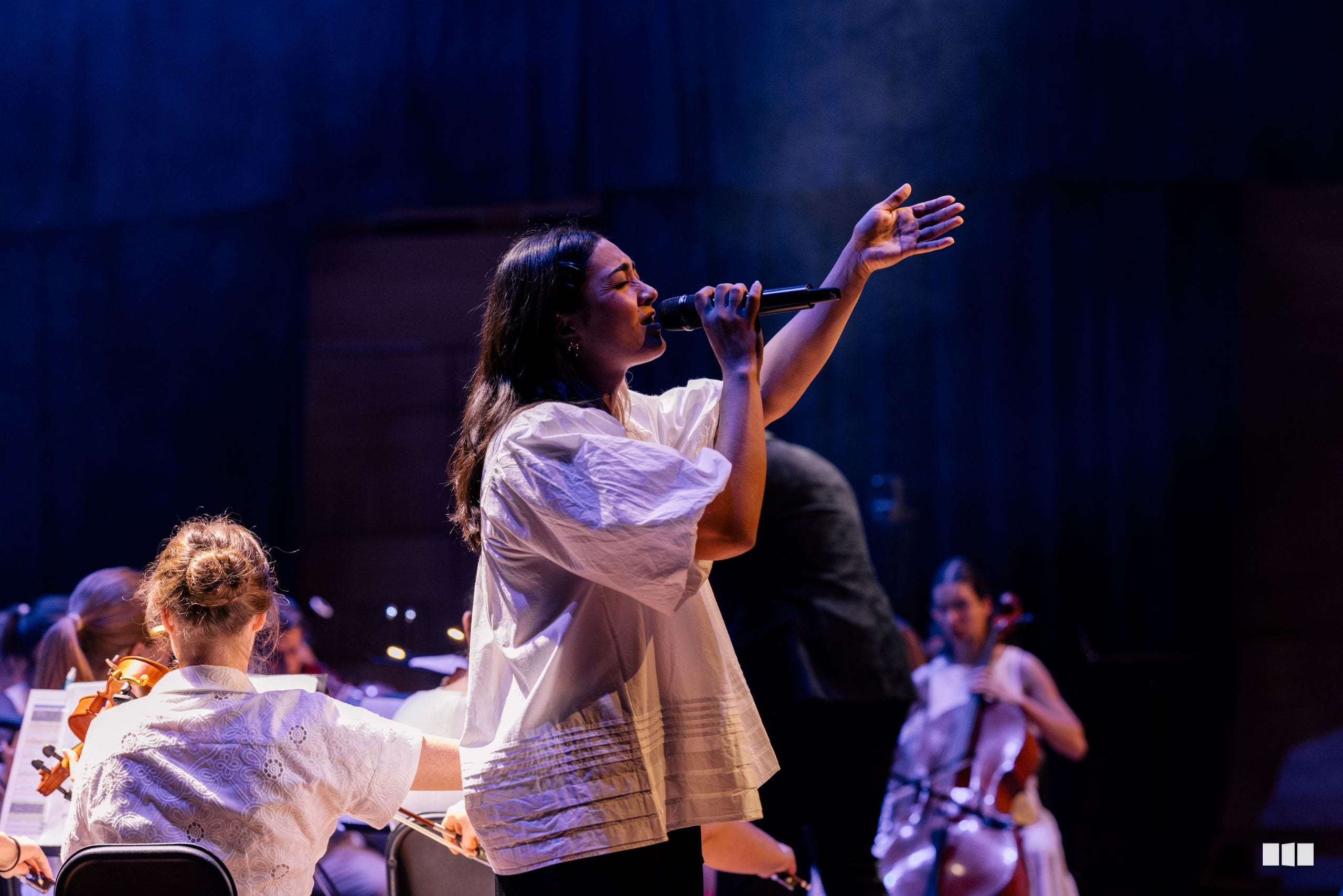 A female soloist performs passionately with microphone while a young child watches from the front row during an orchestral concert.