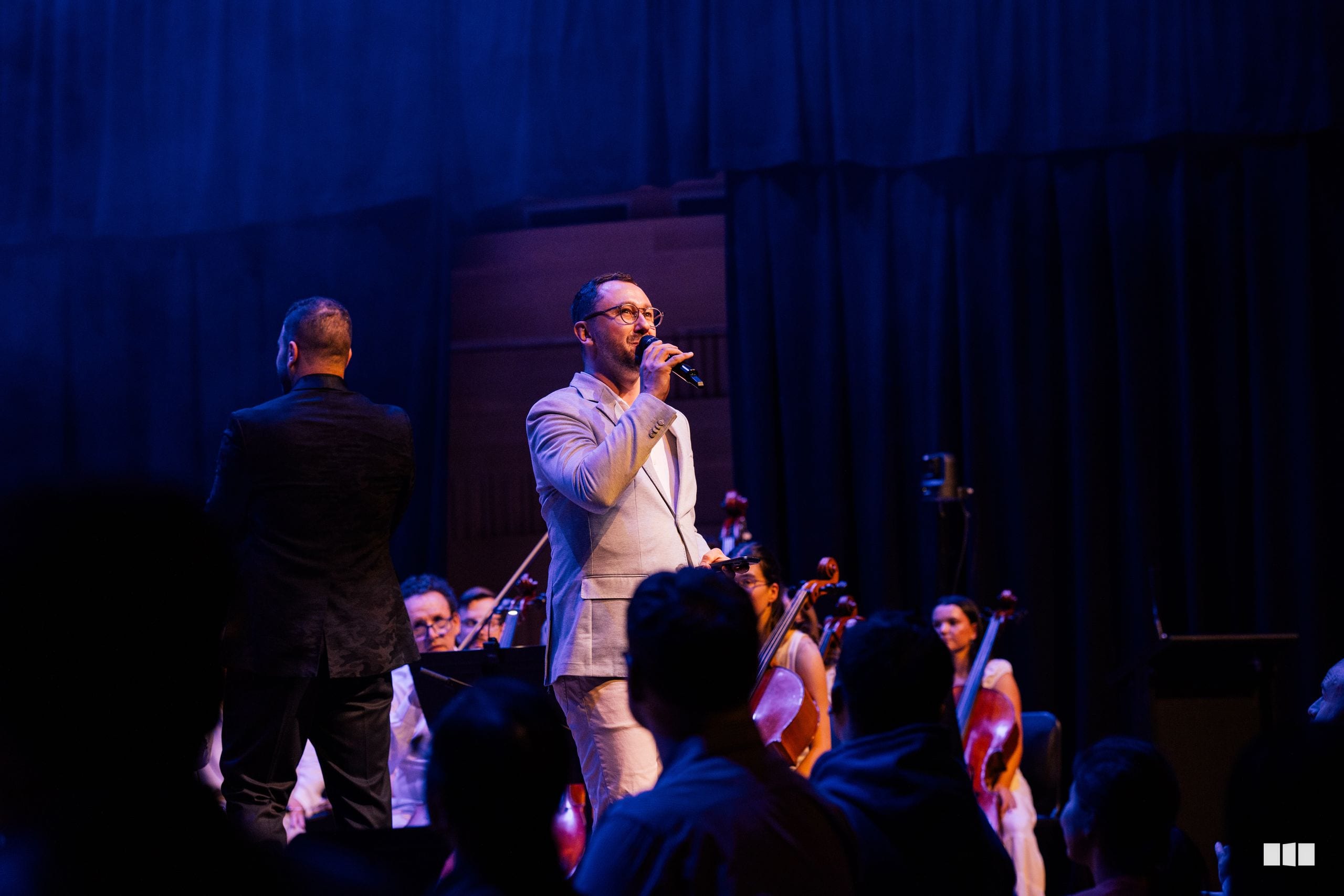 A conductor leads a performance with orchestra members visible in the background during an evening concert.