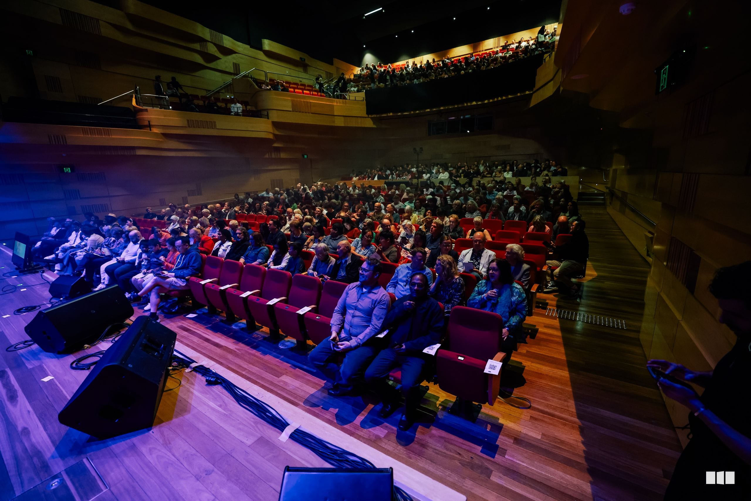 A full auditorium of attendees seated in burgundy chairs watches a performance on stage during an evening event.