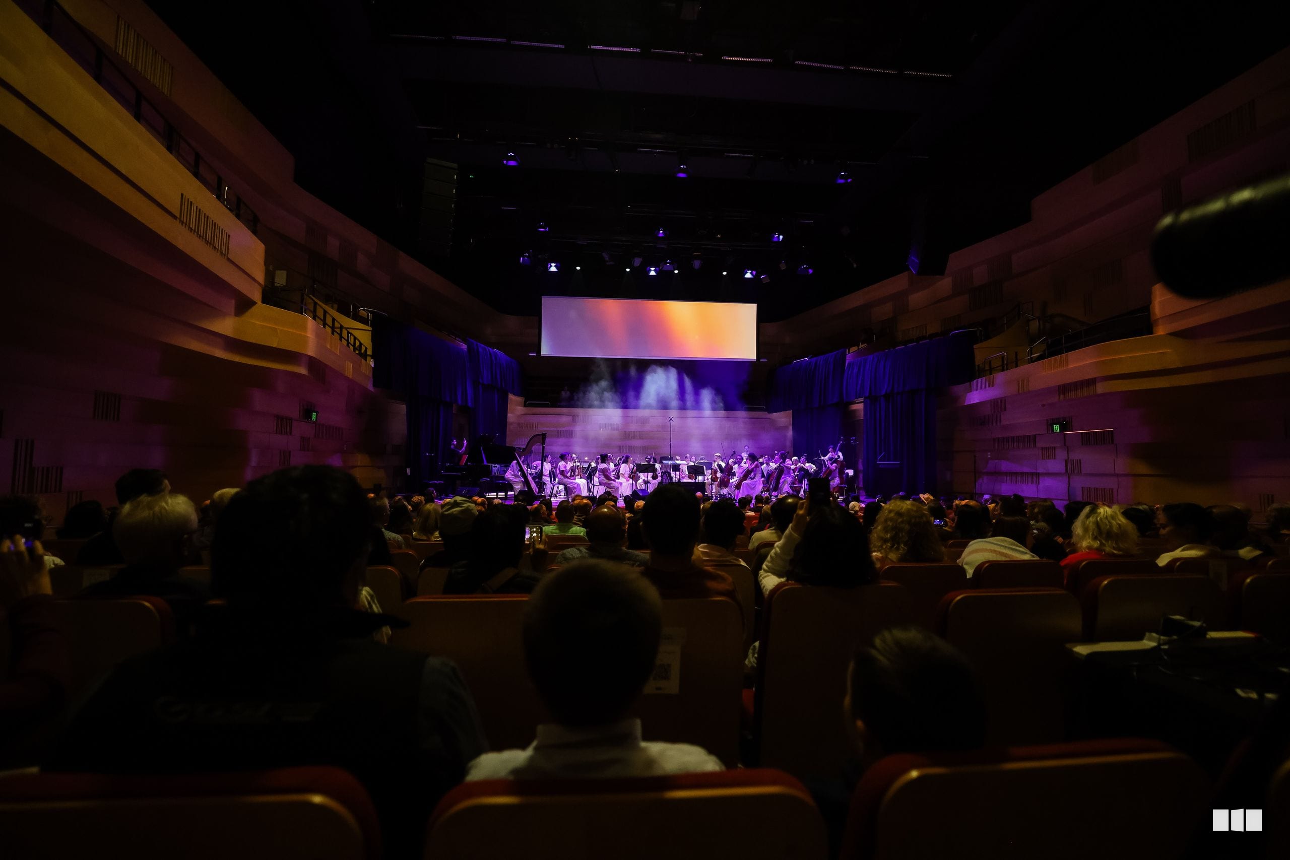 A full orchestra performs on stage beneath atmospheric lighting while an engaged audience watches from the auditorium.