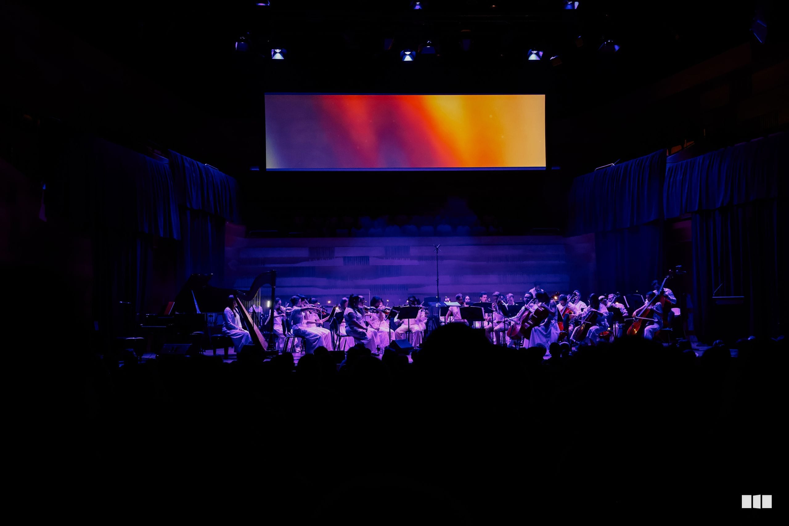 A full orchestra performs on stage beneath a vibrant gradient display screen, with dramatic purple and blue lighting illuminating the musicians and audience in the foreground.
