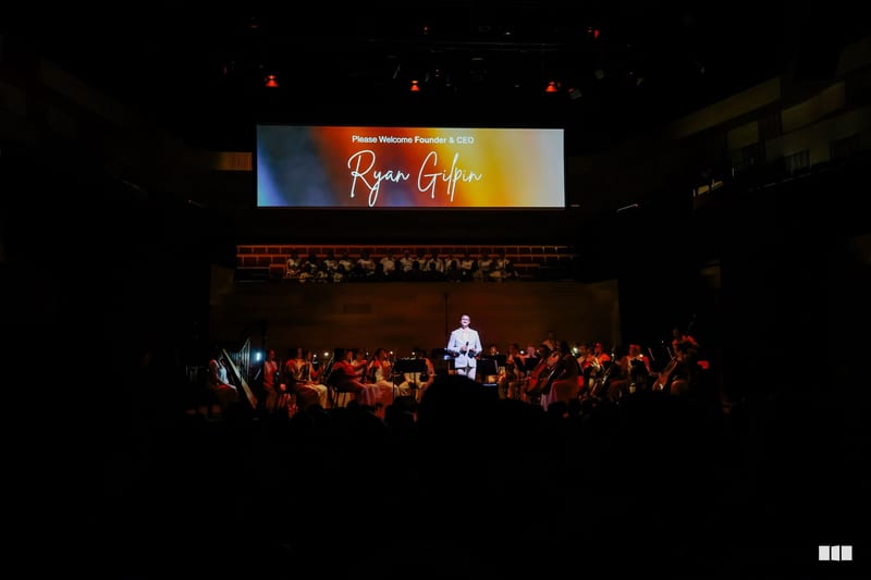 Founder and CEO Ryan Giffin is welcomed to the stage at a formal orchestral event with musicians and audience members present.