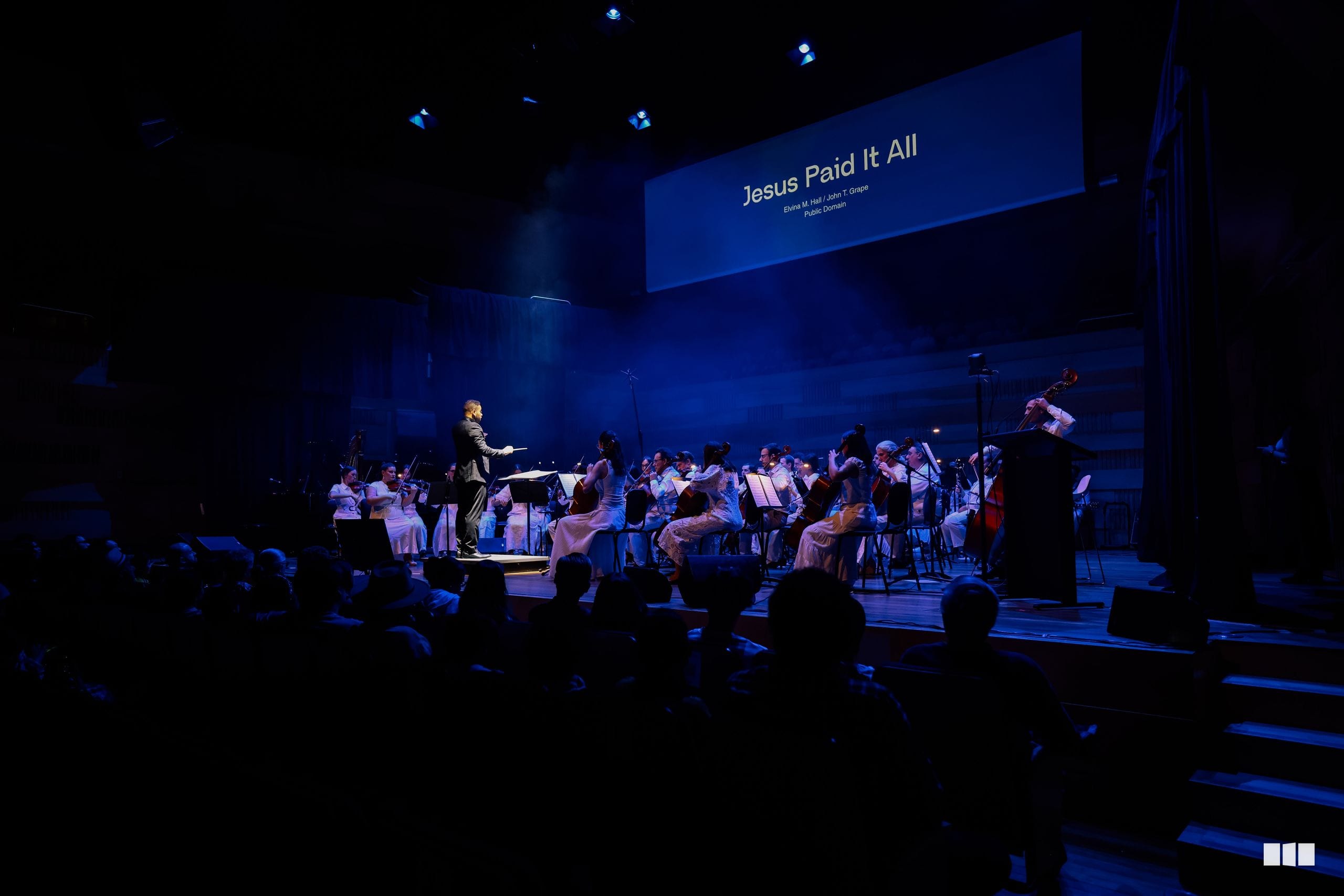 A conductor leads a full orchestra and choir performing beneath a projection reading 'Jesus Paid It All' in a contemporary worship setting.