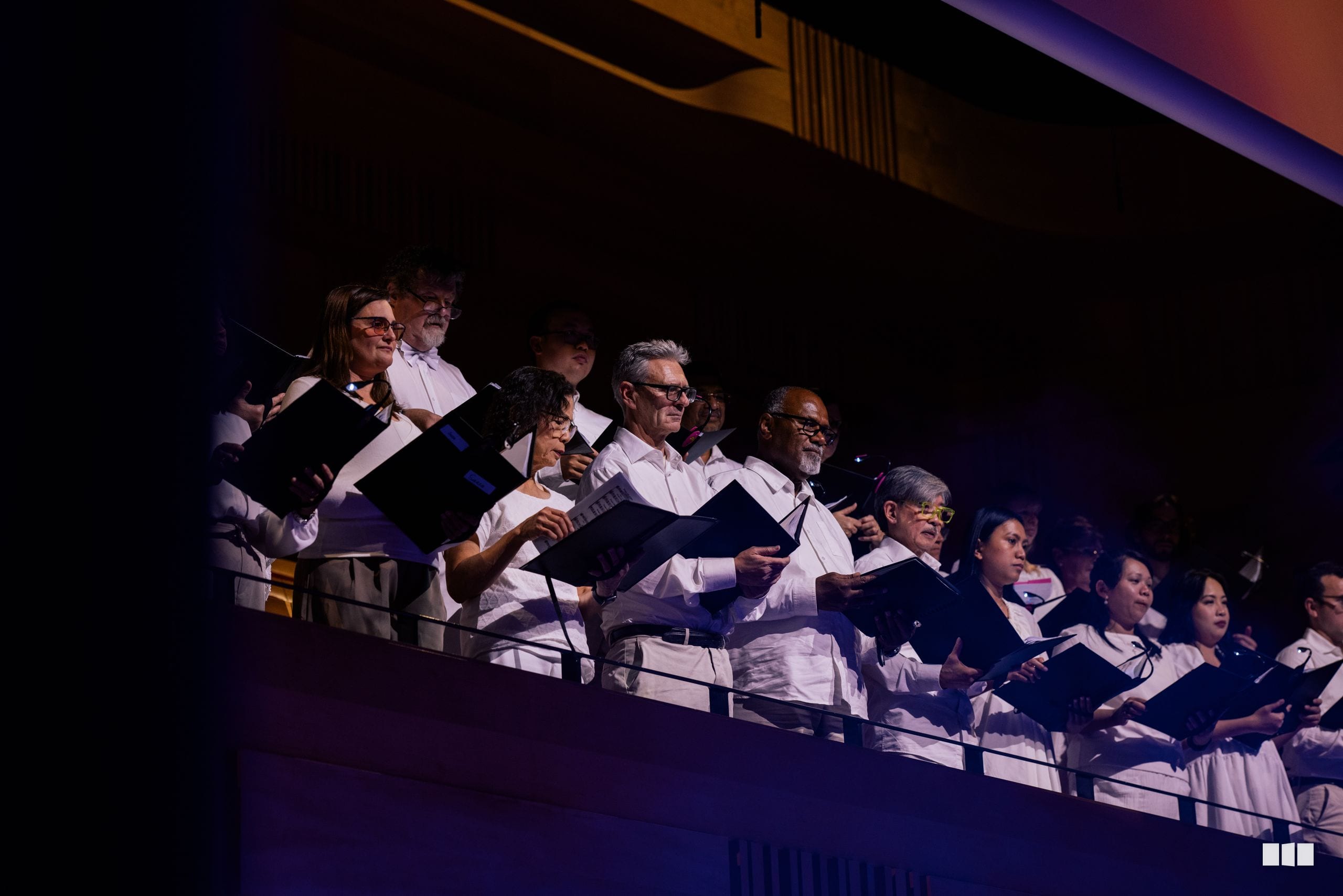 A choir performs on stage in formal attire, holding sheet music and singing together under stage lighting.