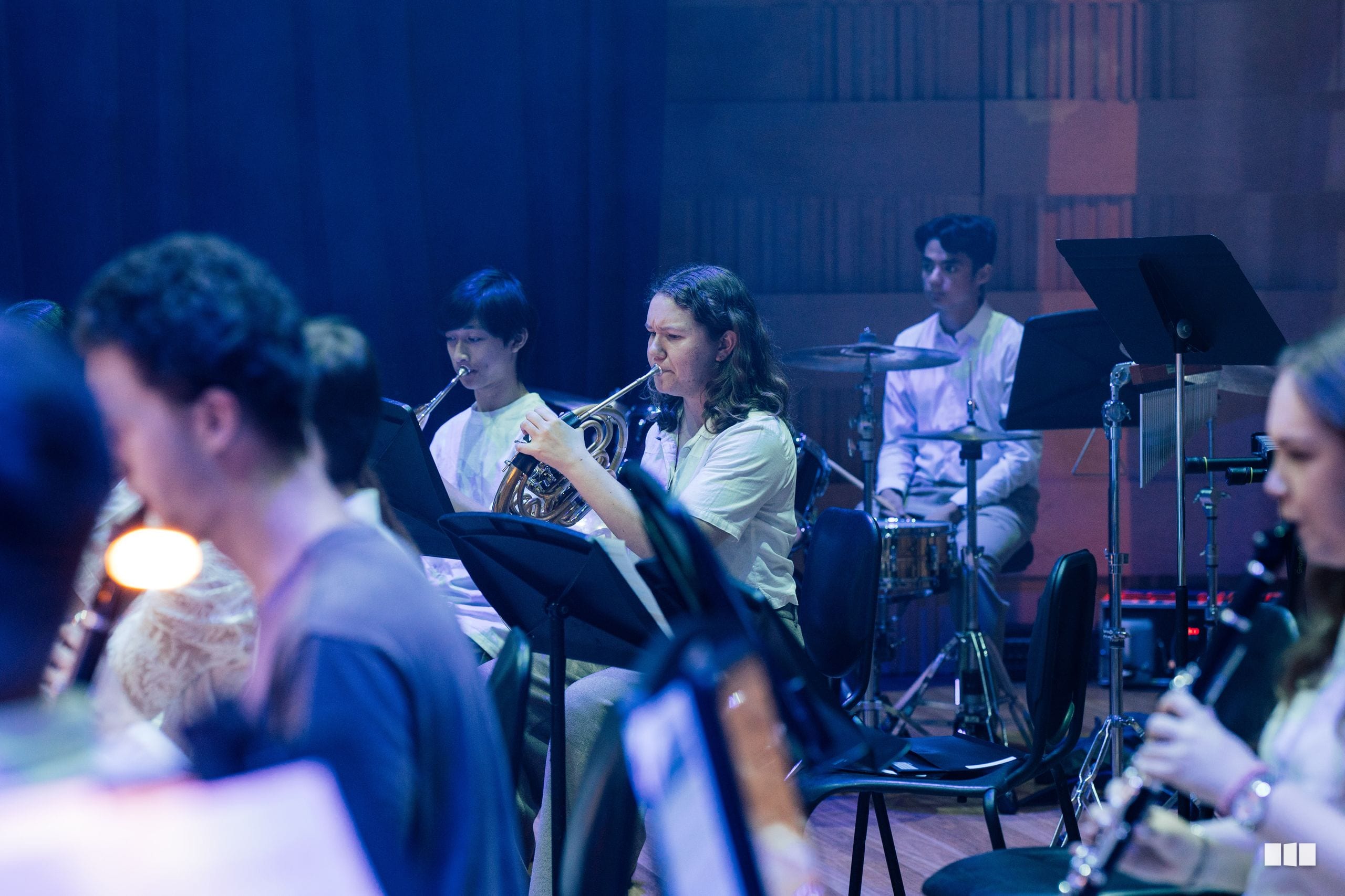 A young musician plays French horn during an orchestral rehearsal or performance with ensemble members and conductor visible on stage.