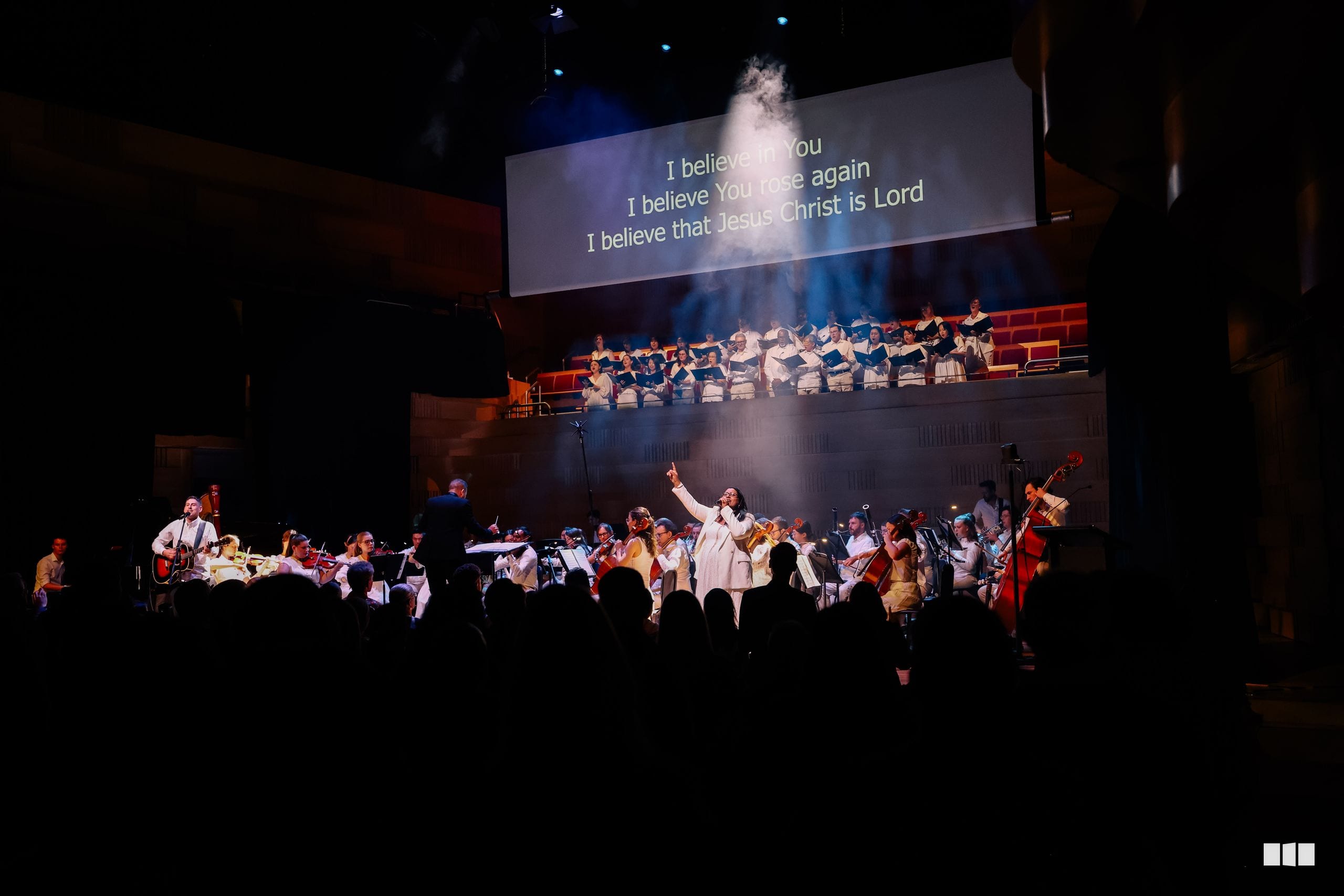 A full orchestra and choir perform together on stage beneath a projection displaying Christian affirmation text, with audience members visible in the foreground.