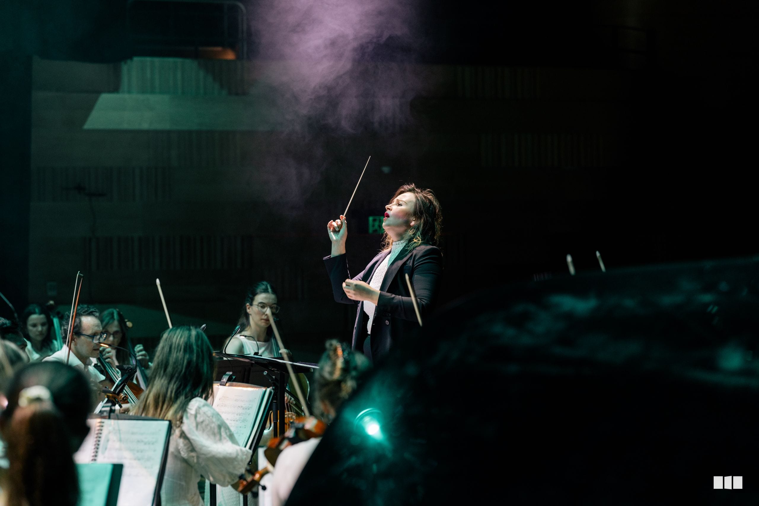 A conductor leads an orchestra during a performance, with string players visible in the foreground.