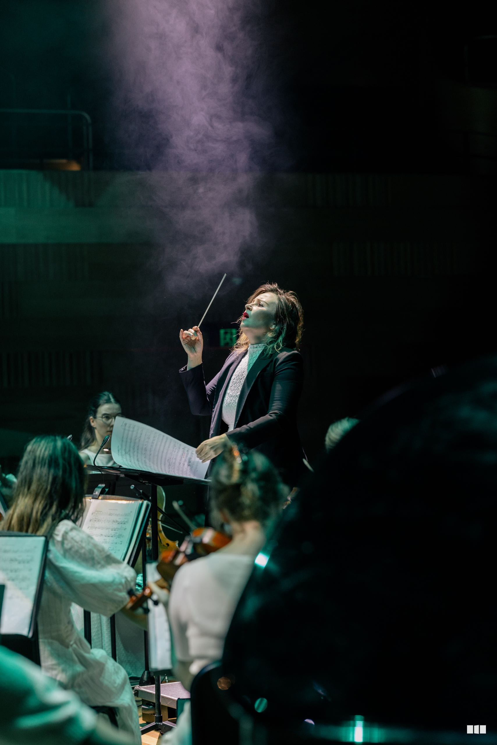 A conductor leads an orchestra during a performance, with dramatic atmospheric lighting illuminating the scene.