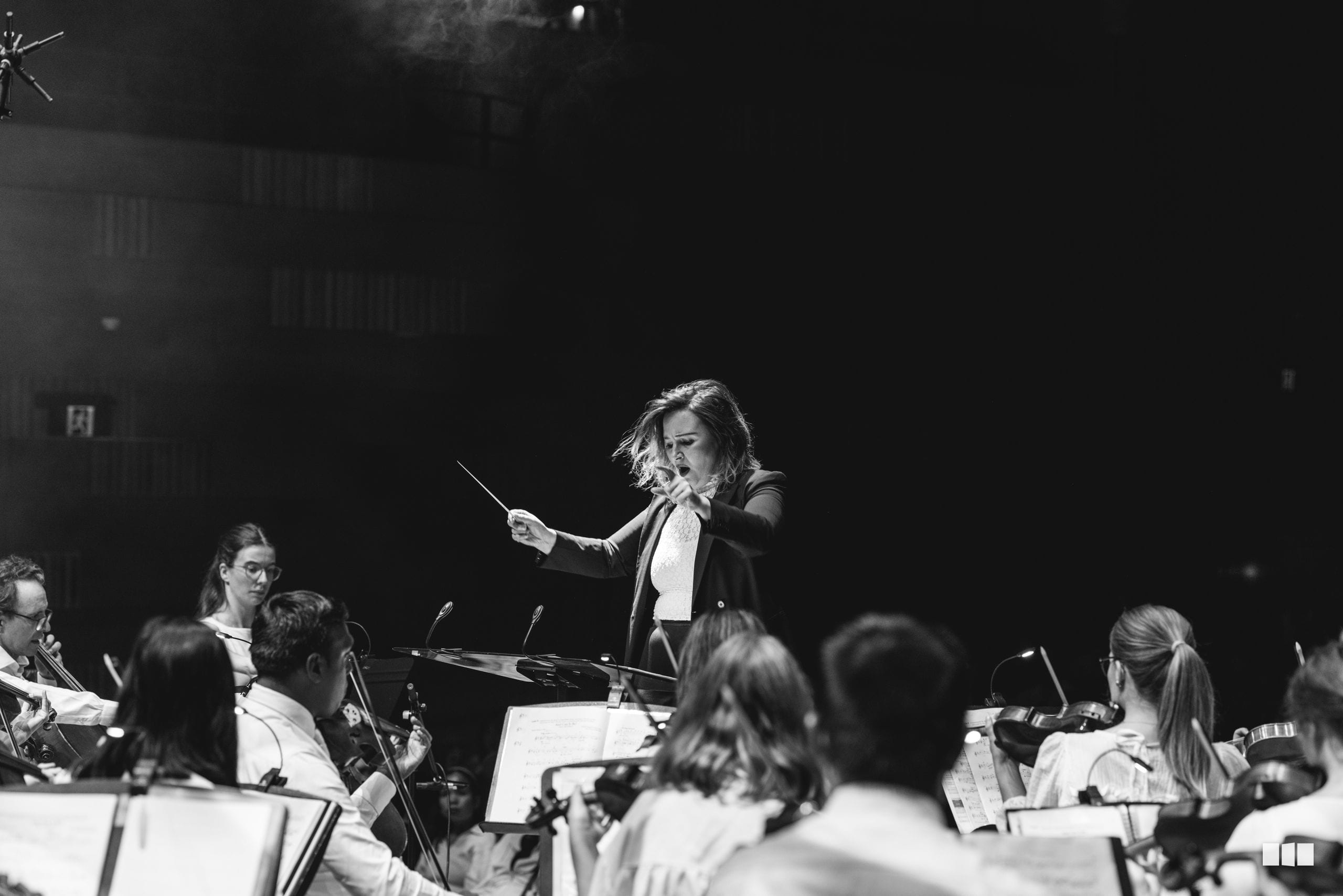 A conductor leads an orchestra during a performance, with string and wind players visible in the ensemble.
