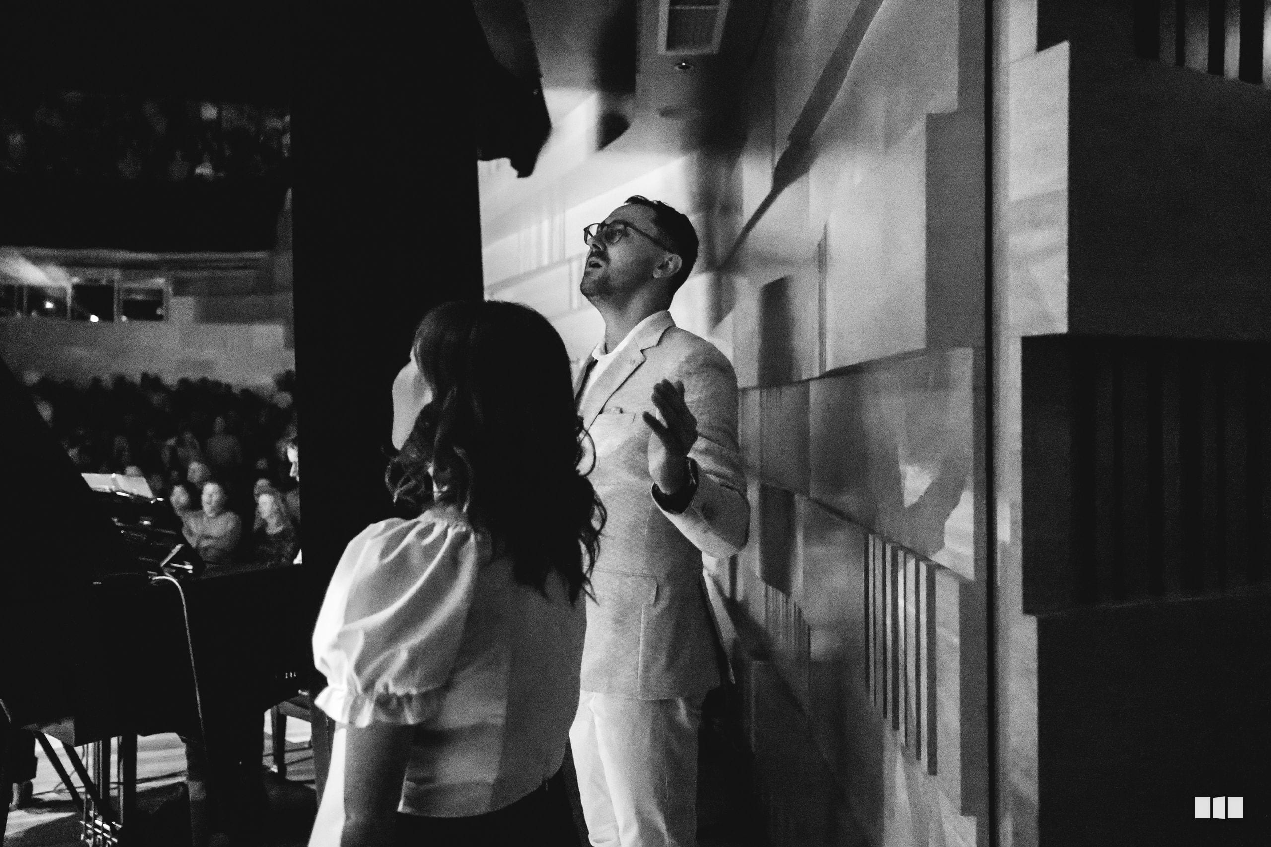 A man in glasses and light-colored shirt gestures expressively while speaking to a woman at an outdoor evening event.