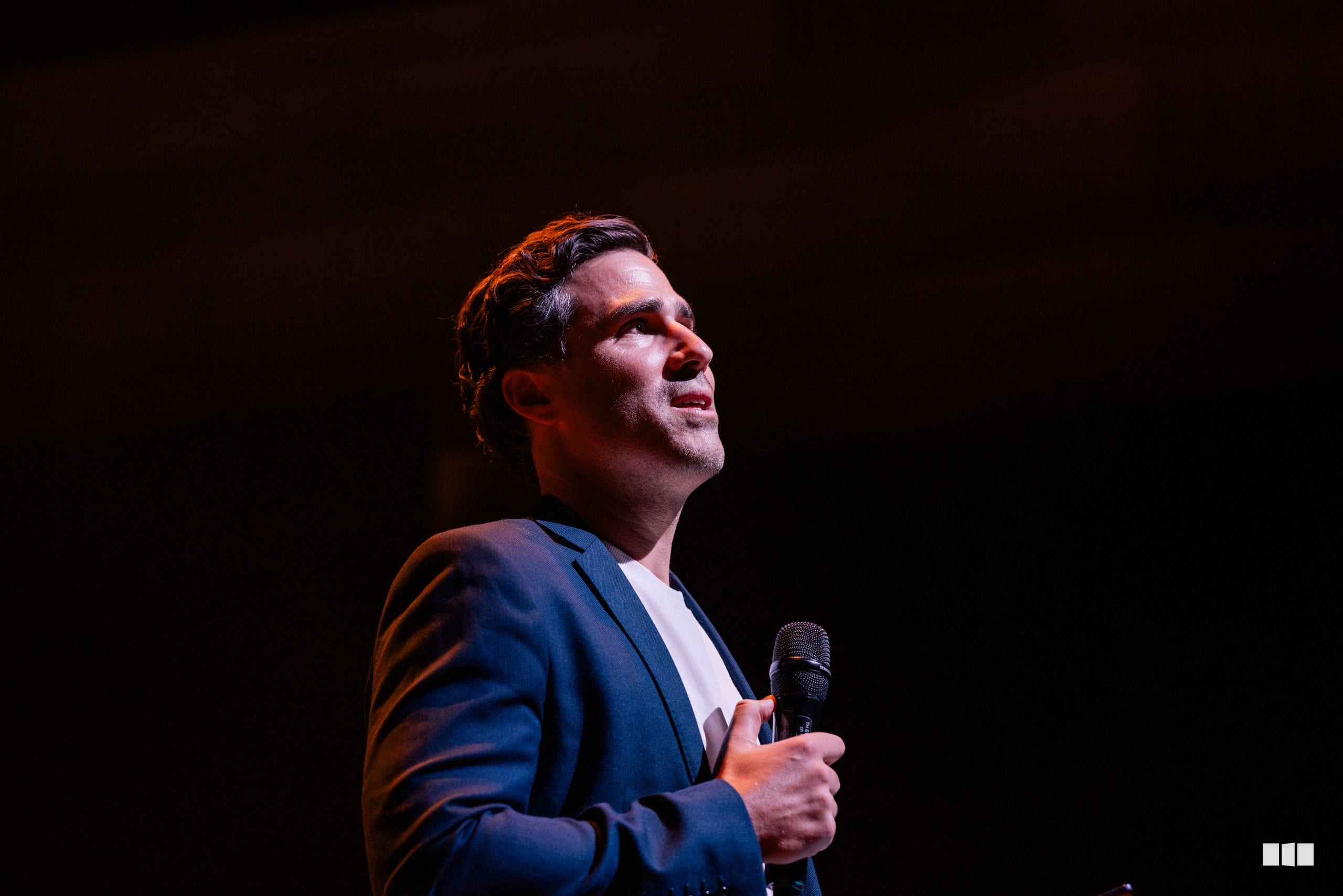 A man in a navy blazer and white shirt holds a microphone while speaking on stage against a dark background.