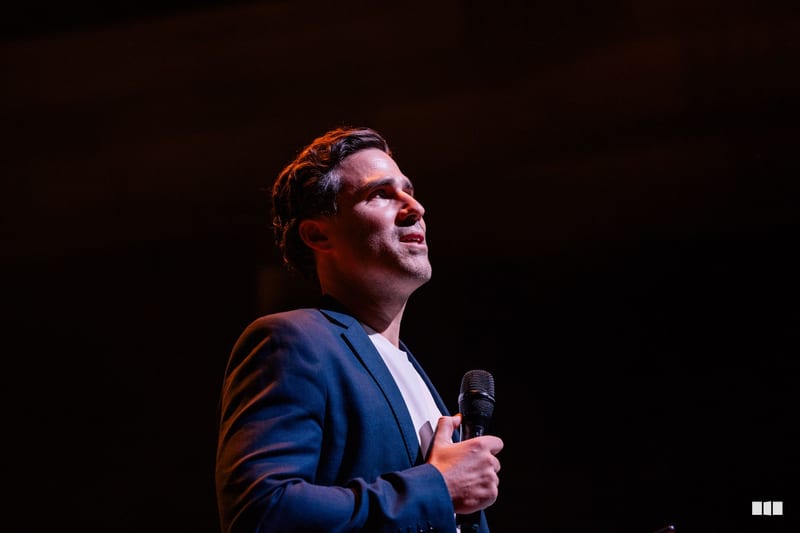 A man in a navy blazer and white shirt holds a microphone while speaking on stage against a dark background.