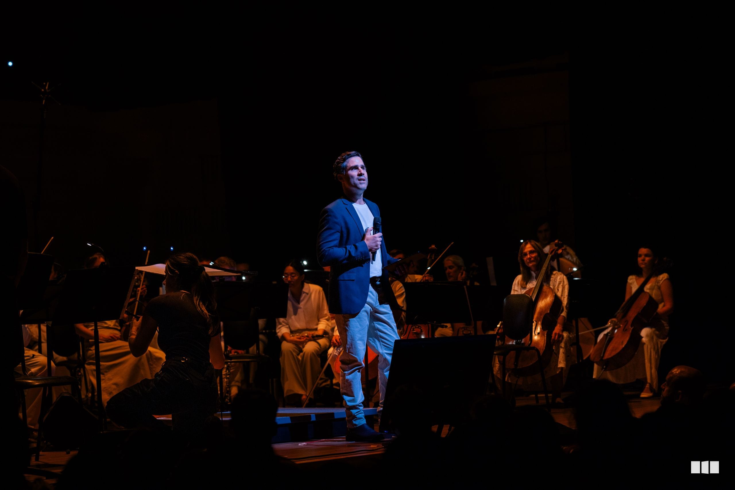 A male soloist in a blue blazer performs with a string orchestra on stage under warm spotlight.