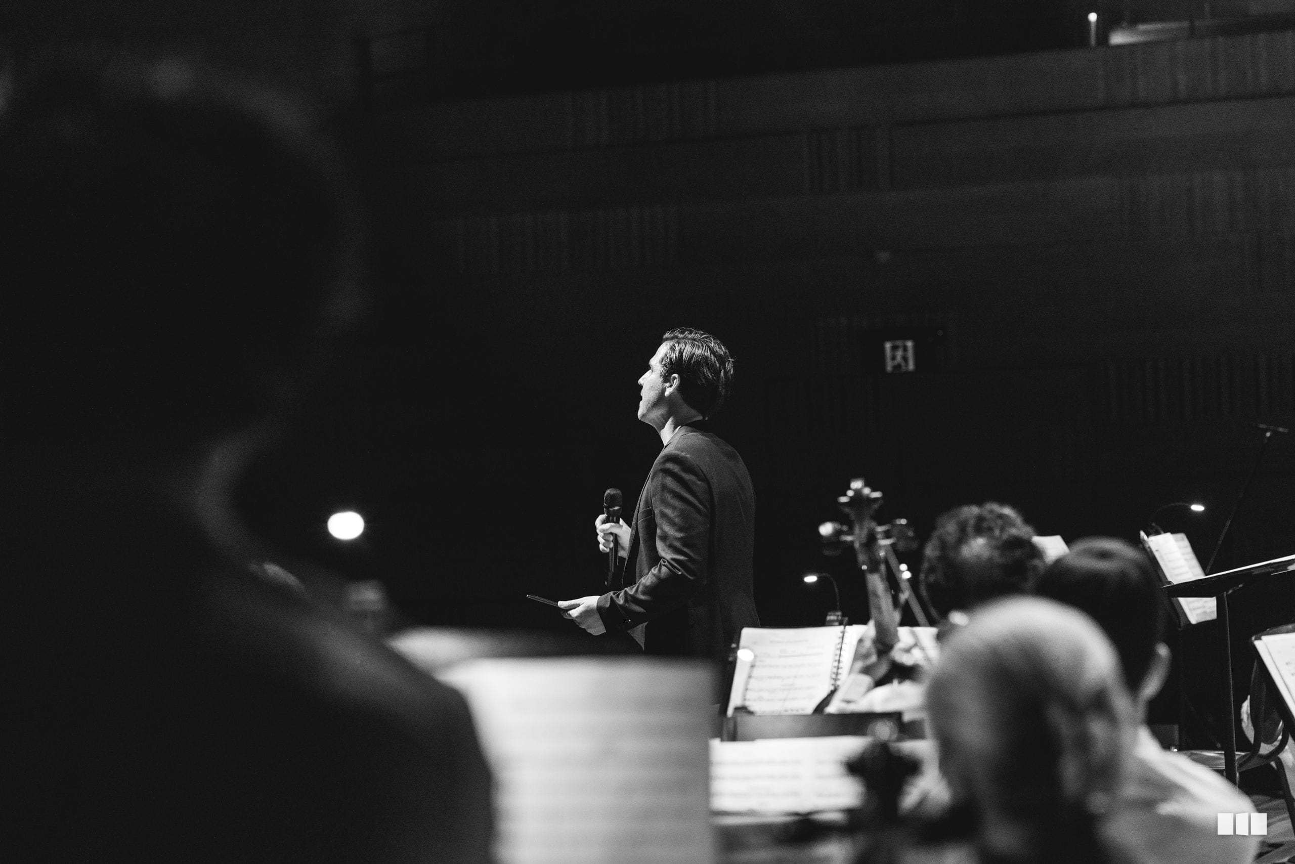 A conductor leads an orchestra during a performance in a concert hall, with musicians and sheet music visible in the foreground.