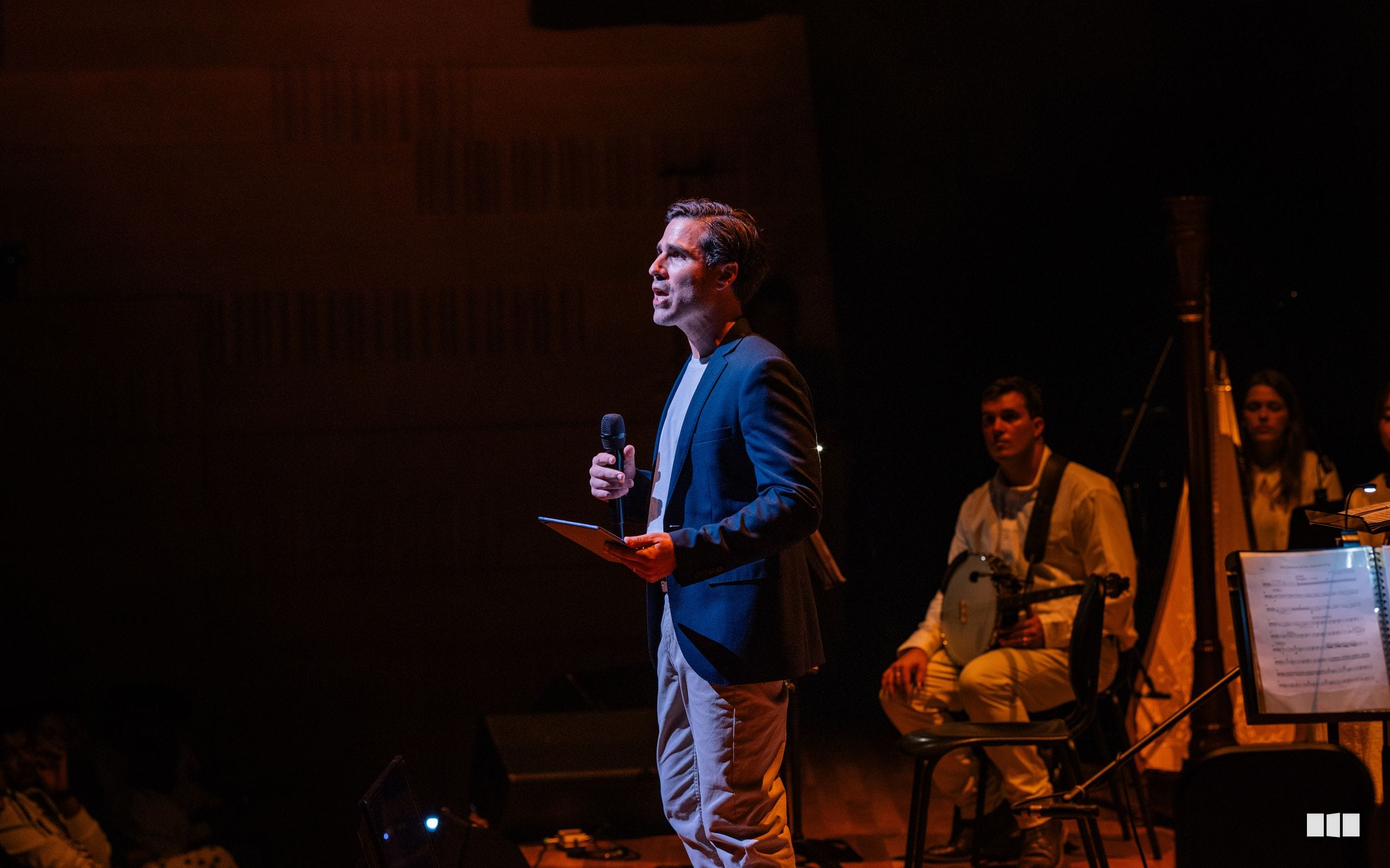 A speaker in a blue blazer addresses the audience while musicians with sheet music sit attentively on stage.