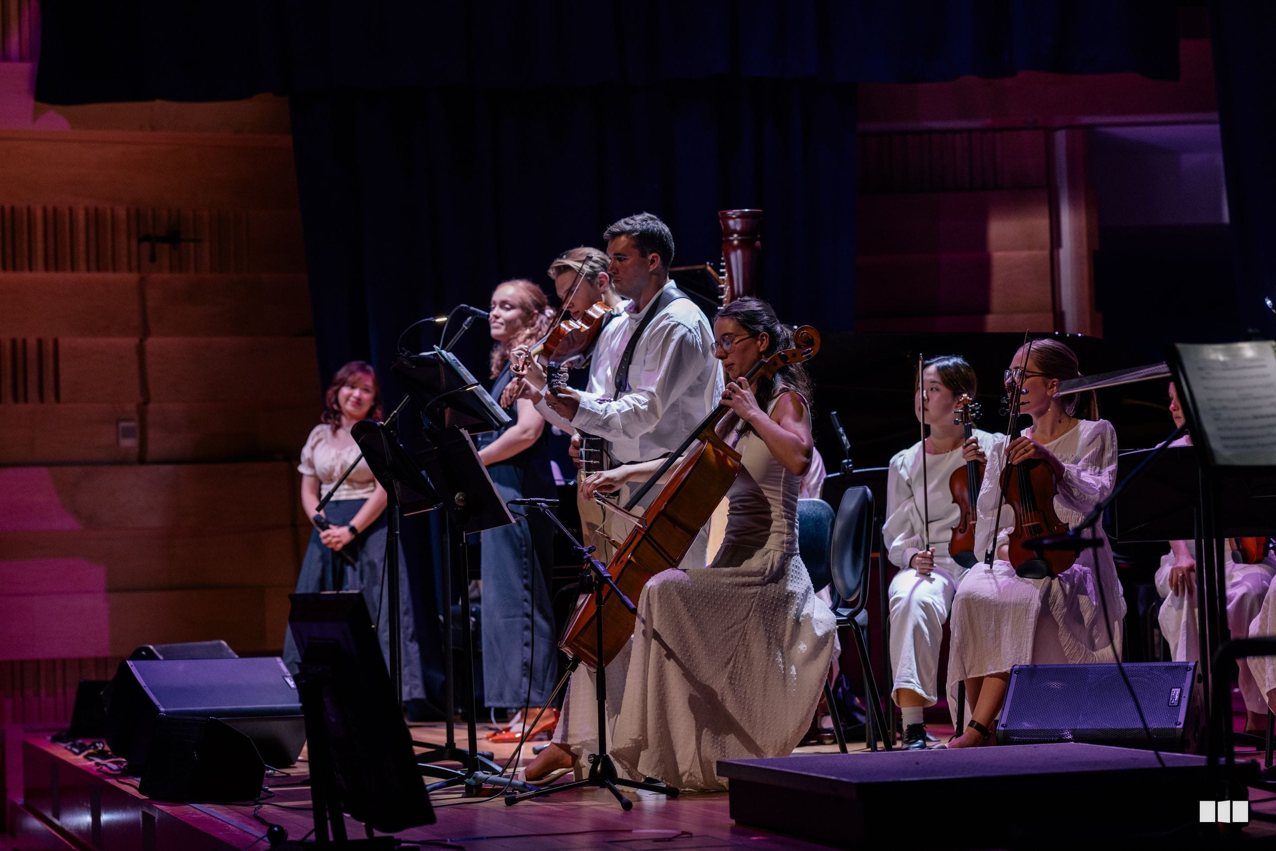A cellist performs as a soloist with orchestral accompaniment on stage under warm stage lighting.