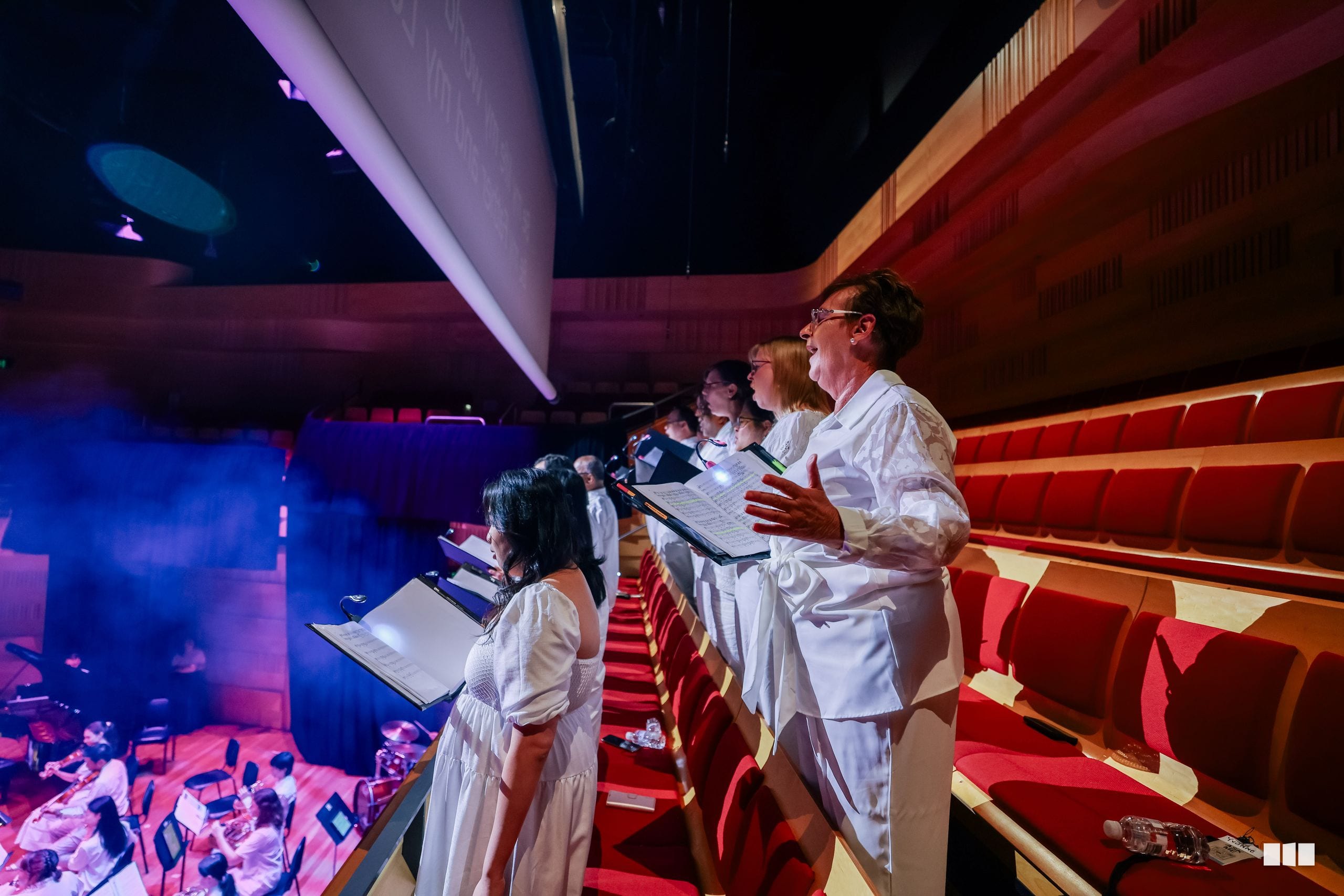Choral singers in white robes perform with sheet music on stage while an orchestra plays below under atmospheric purple and red lighting.