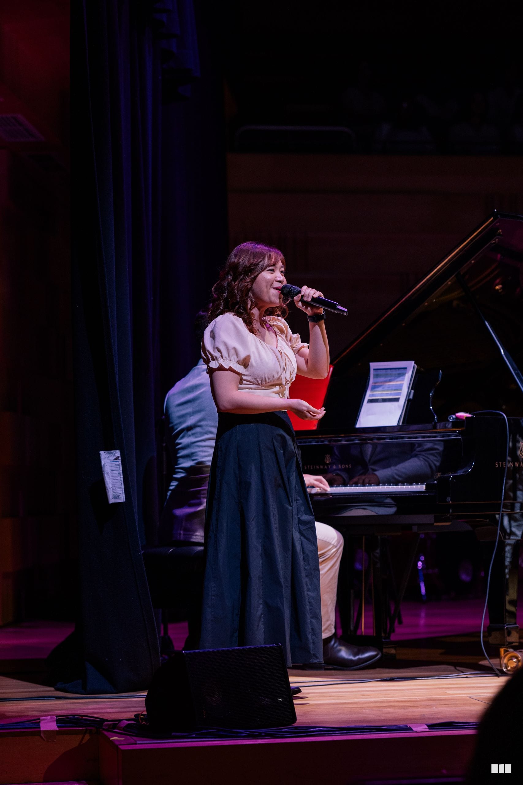 A female soloist performs at the microphone with a grand piano and laptop visible on stage during an evening concert.