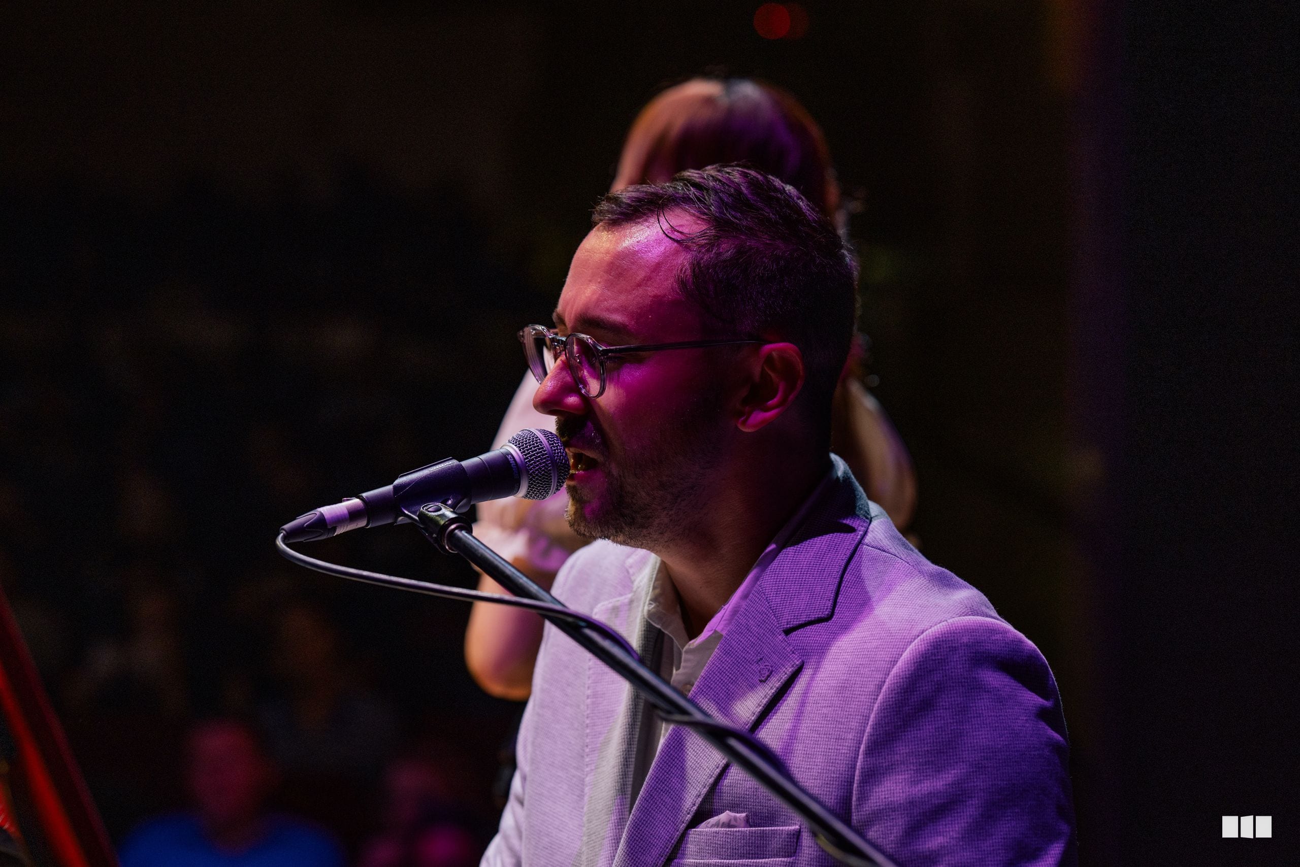 A male soloist wearing glasses and a purple jacket sings into a microphone at the stage.