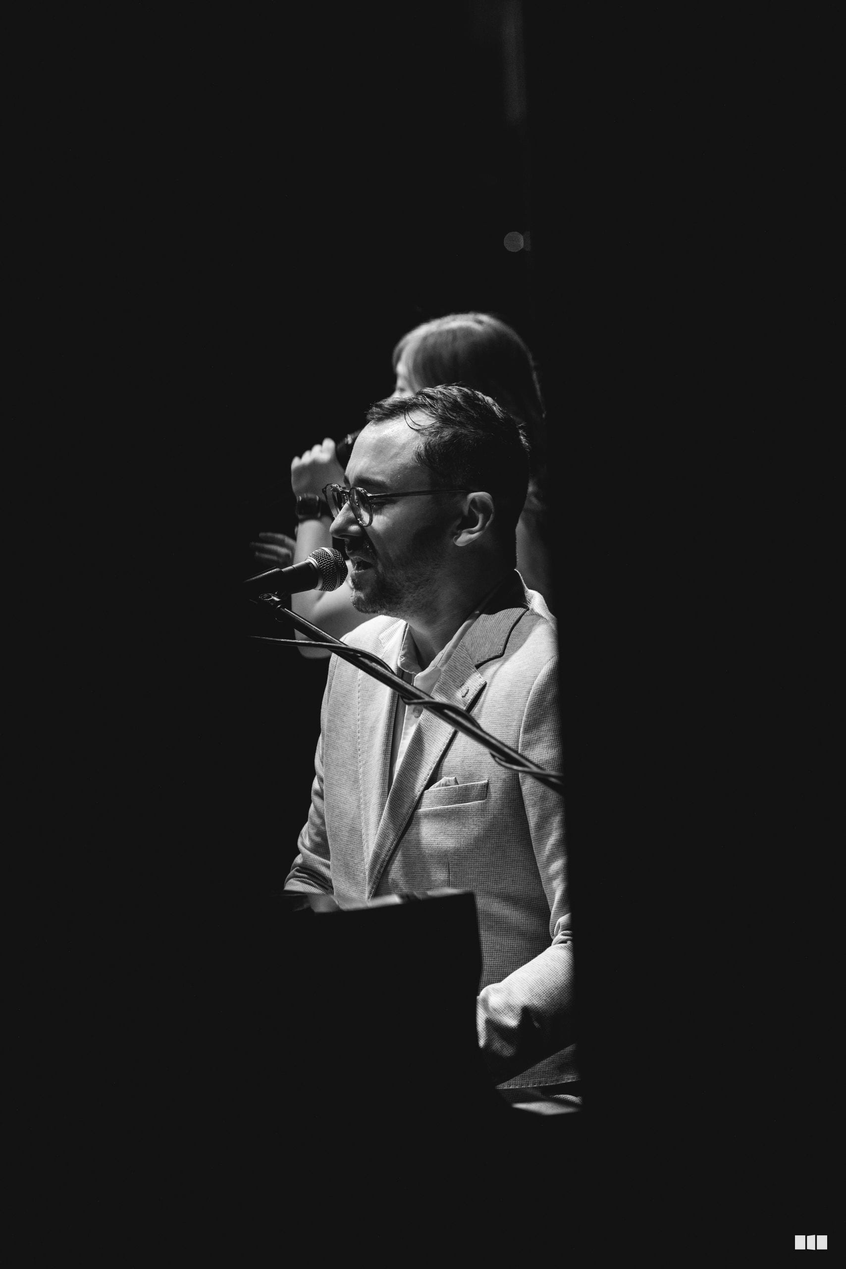 A male soloist wearing glasses and a light-colored blazer performs at the microphone against a dark background.