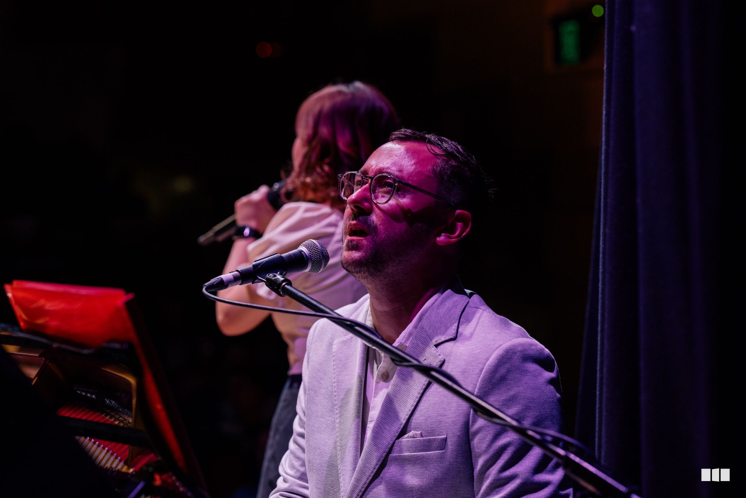 A male performer wearing glasses and a light purple blazer sings into a microphone at the piano during an evening performance.