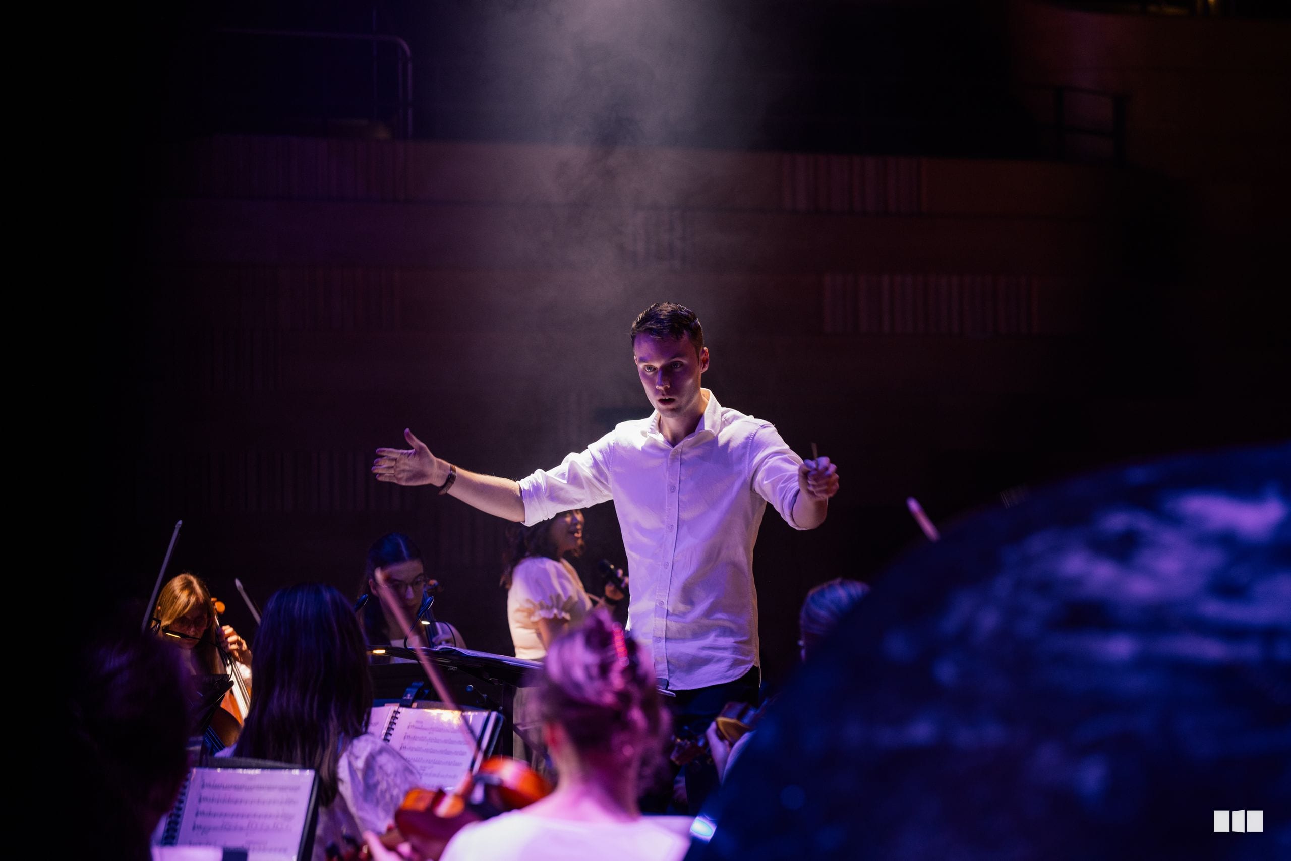 A conductor leads an orchestra with expressive gestures during a concert performance under atmospheric stage lighting.