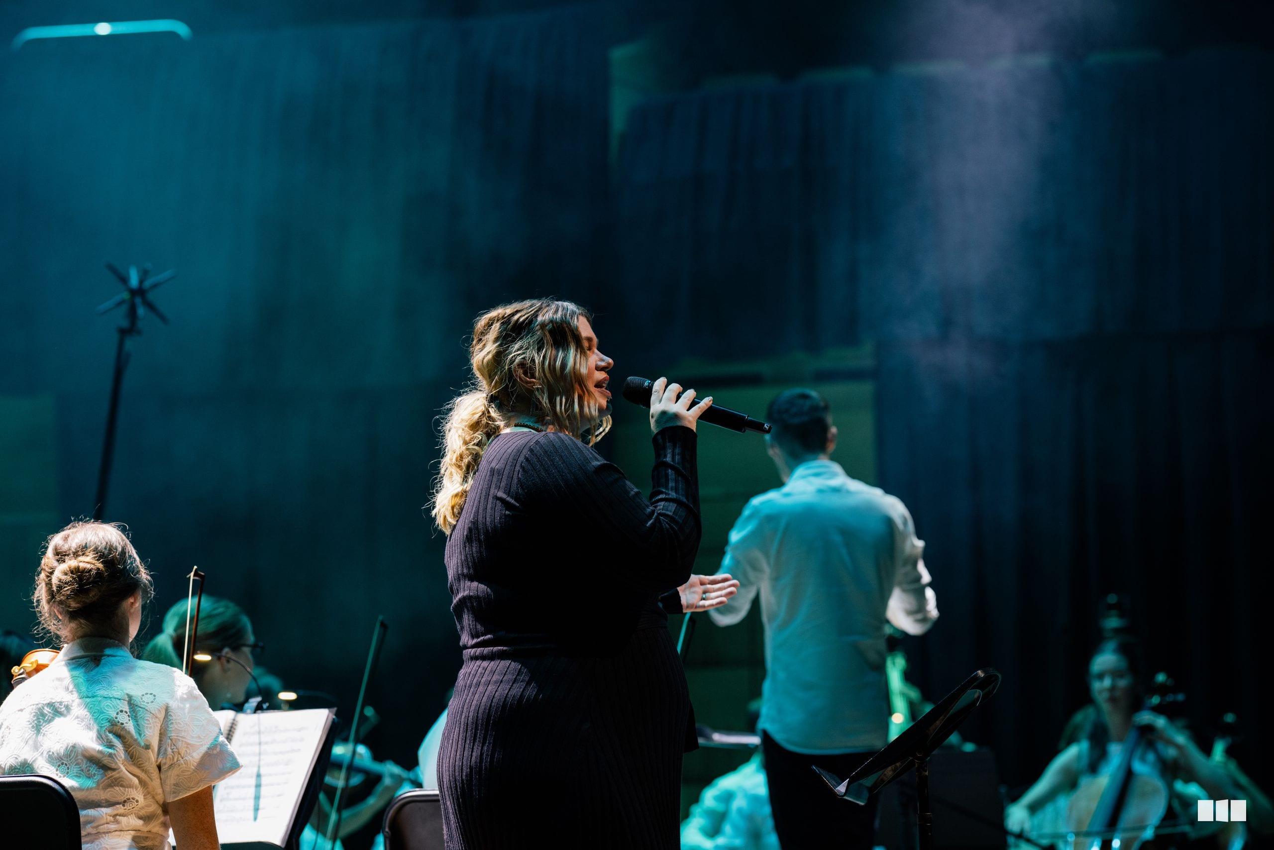 A female soloist performs at the microphone alongside orchestra and choir members in a concert setting with dramatic blue-green stage lighting.