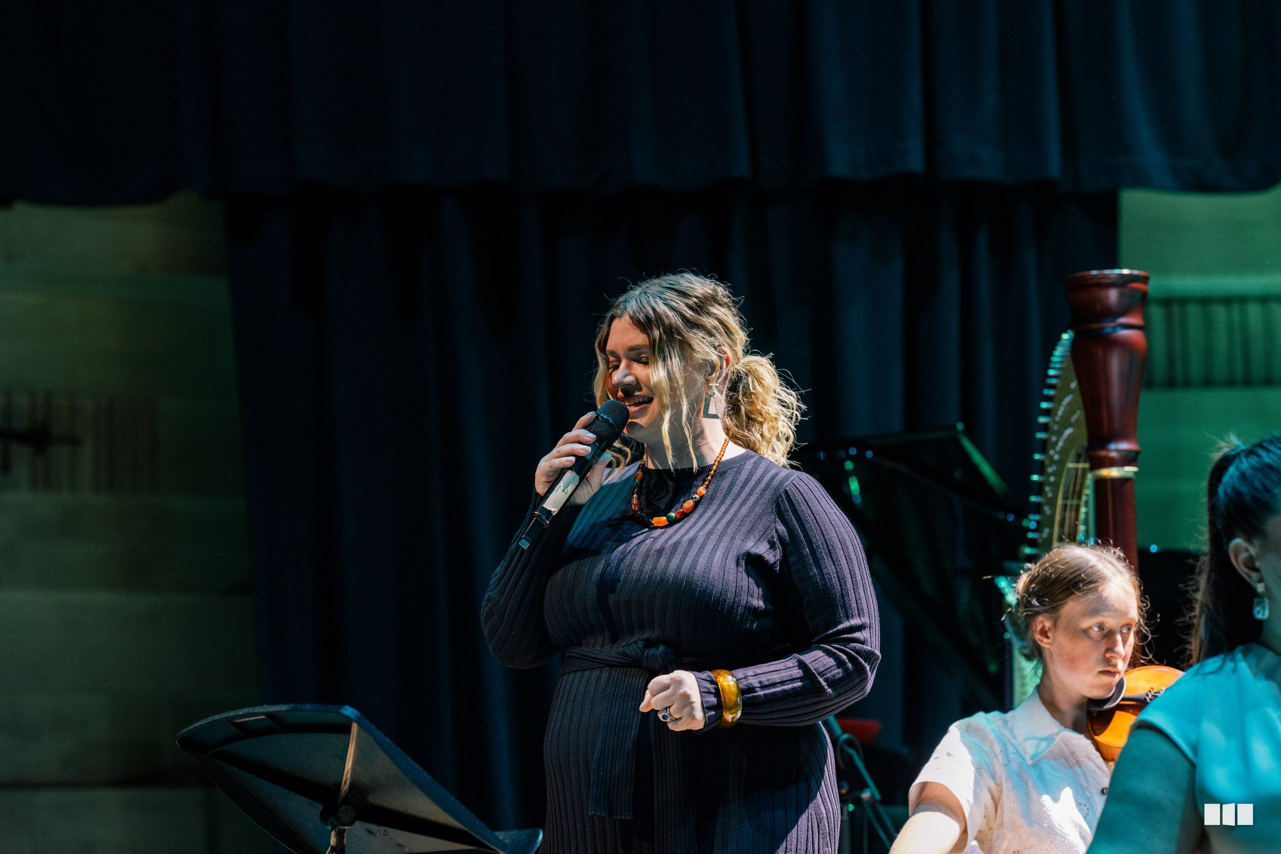 A female soloist in a dark pinstriped dress performs at the microphone on stage with young musicians visible beside her.