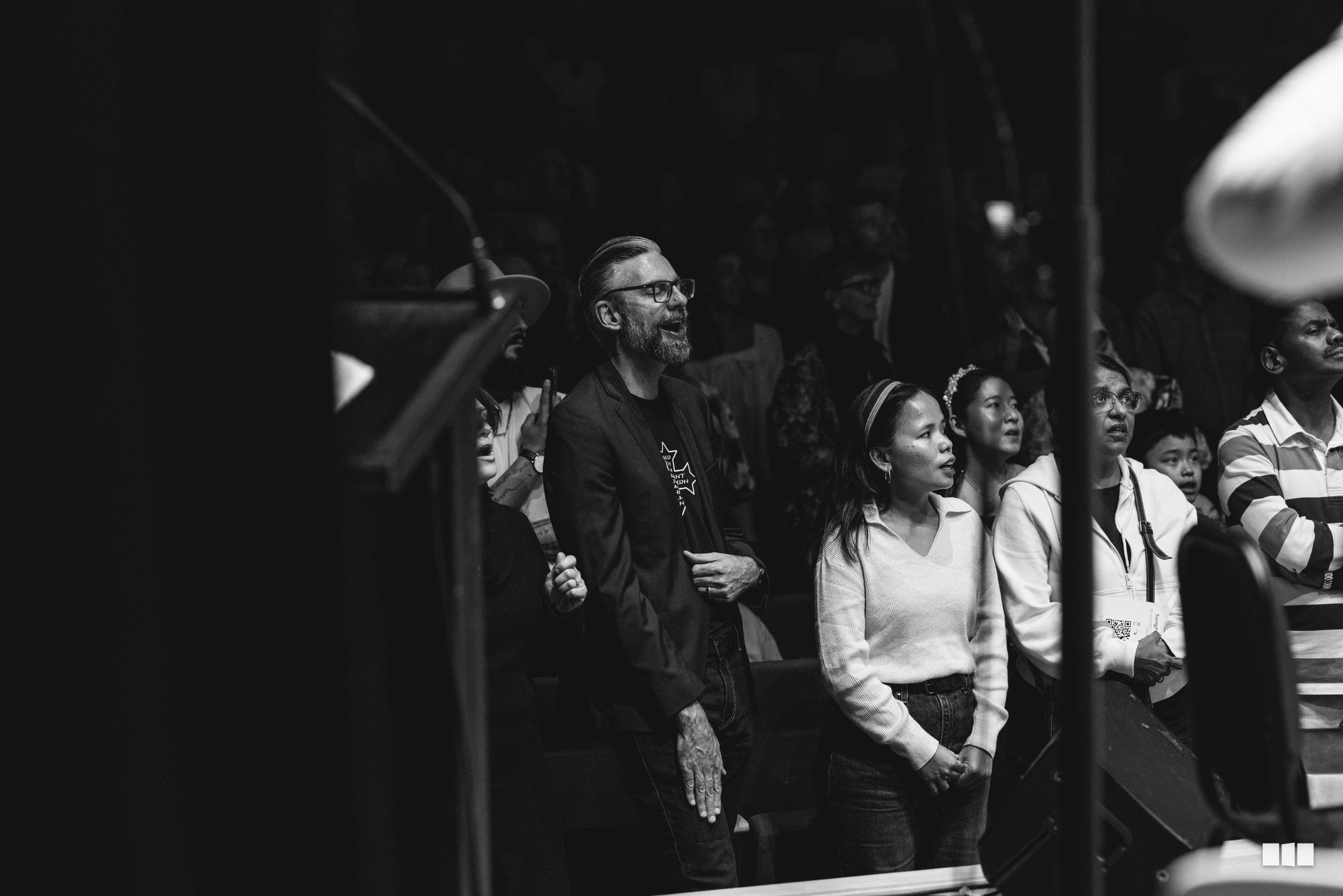 A diverse group of people stand together in an indoor venue, looking upward with engaged expressions during what appears to be a performance or presentation.