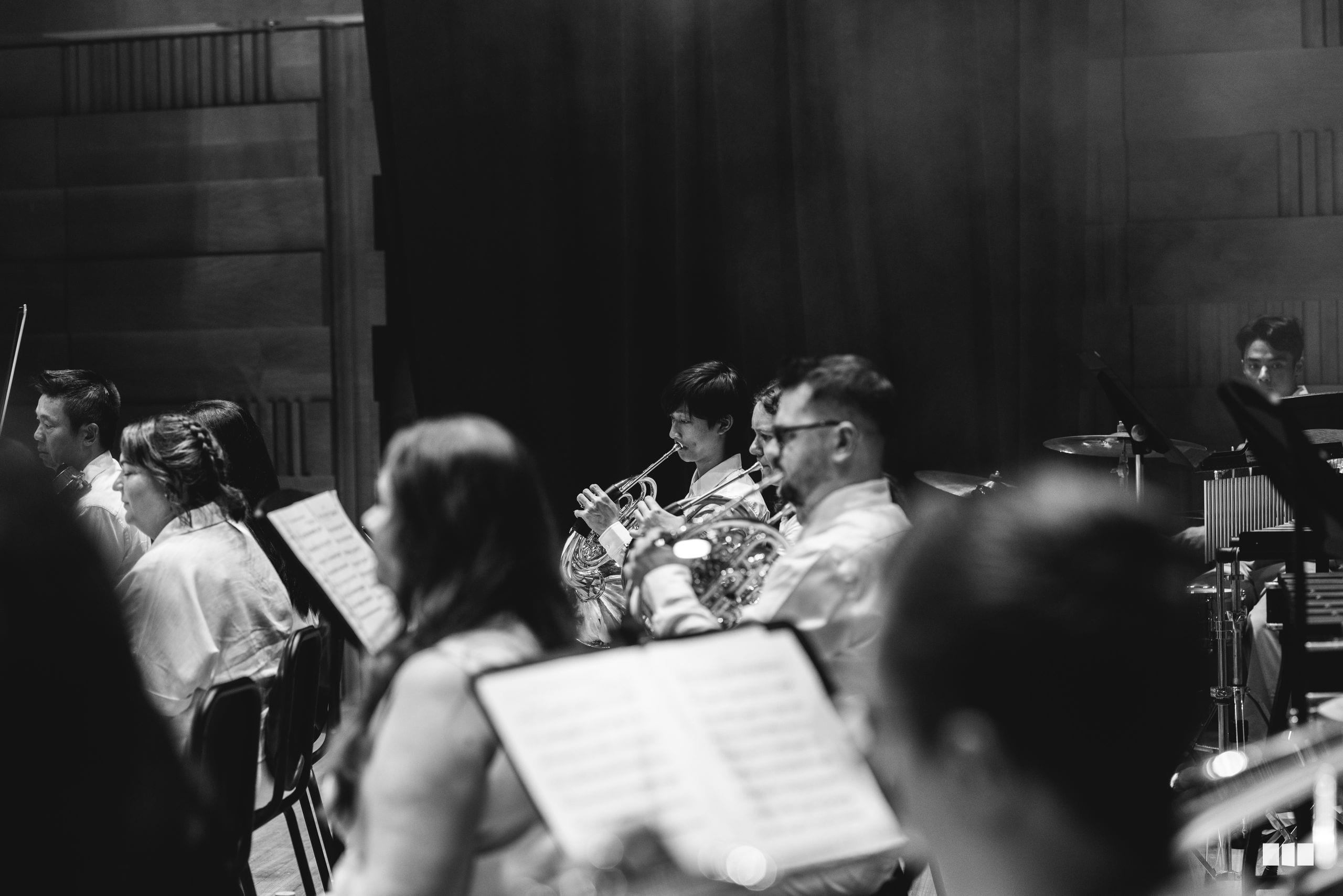 Orchestra musicians perform together during a rehearsal or recording session, with brass and woodwind players visible among the ensemble.