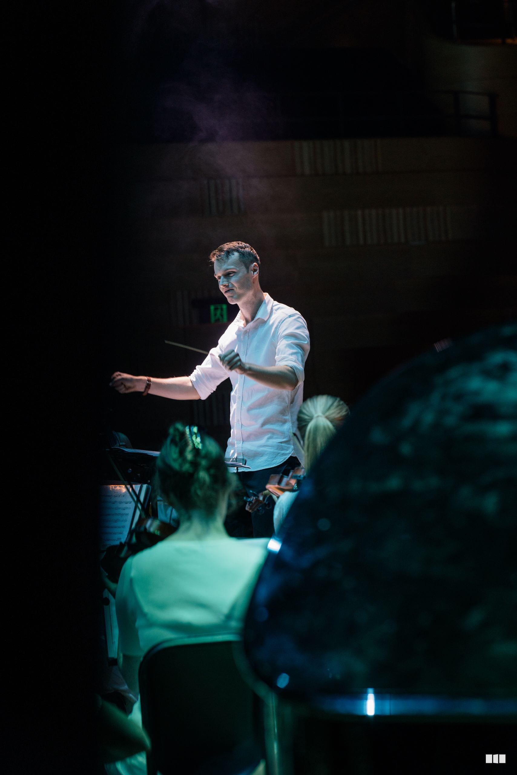 A conductor leads an orchestra during a performance, illuminated by stage lighting with audience members visible in the foreground.